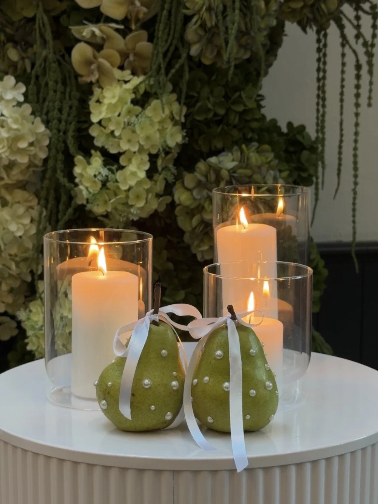 Decorative scene with four lit candles in glass holders and two decorated pears with white ribbons and pearl accents, all placed on a round white surface, with lush green floral arrangements in the background.