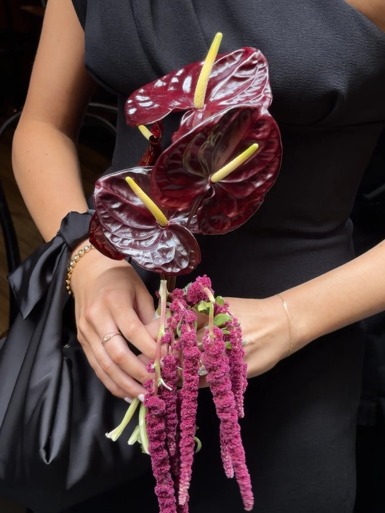 A person holding a bouquet of flowers with dark red anthuriums and bright pink amaranthus, dressed in black clothing.