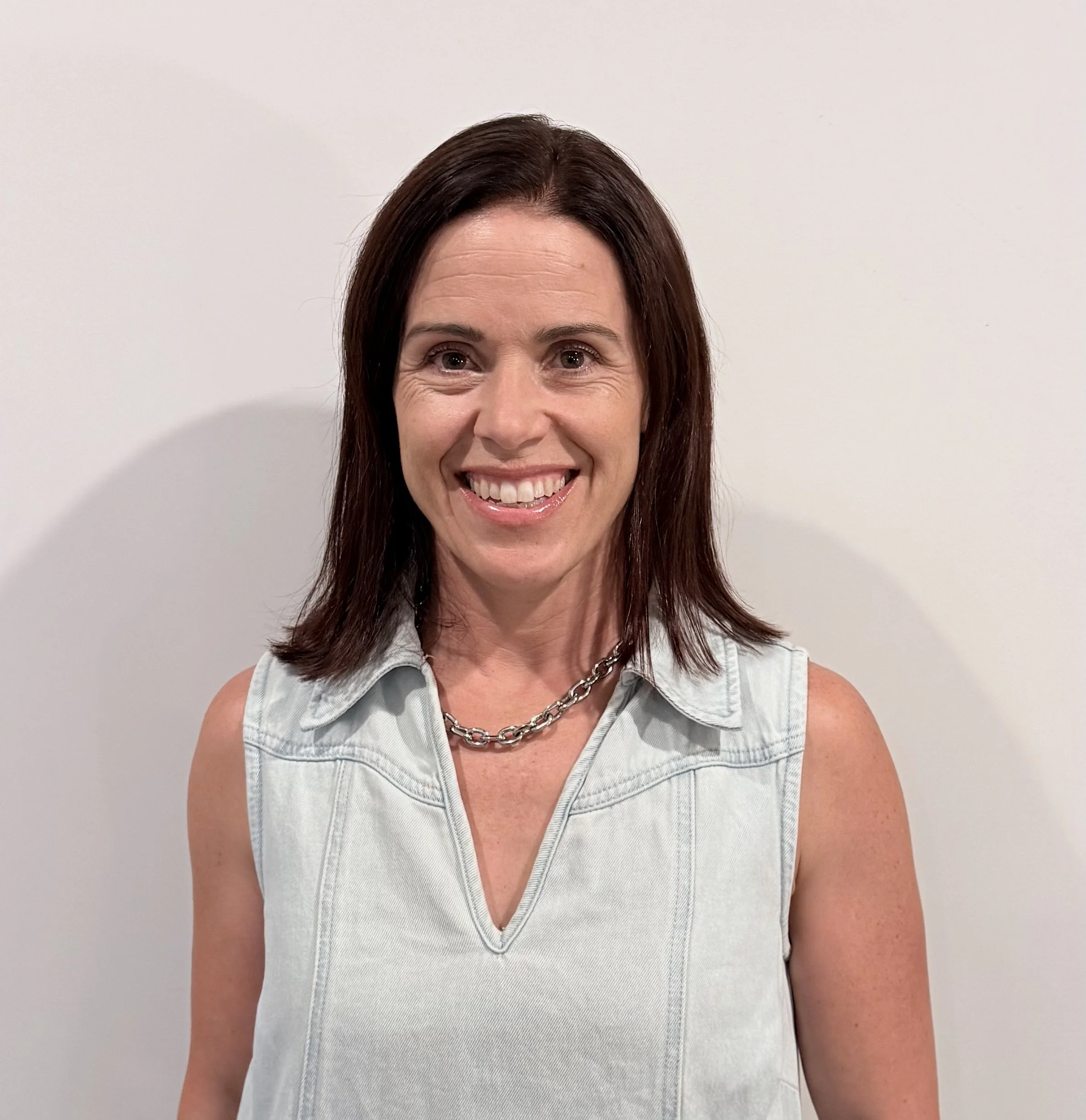 Close-up of a smiling woman with shoulder-length brown hair, wearing a sleeveless denim vest and a chunky chain necklace, standing against a white wall.