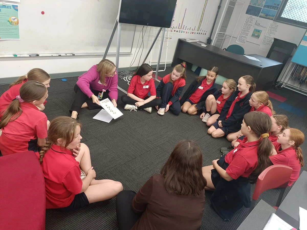 A group of students sitting in a circle on a classroom carpet, listening to a teacher who is showing a poster, with educational posters and a whiteboard on the walls.