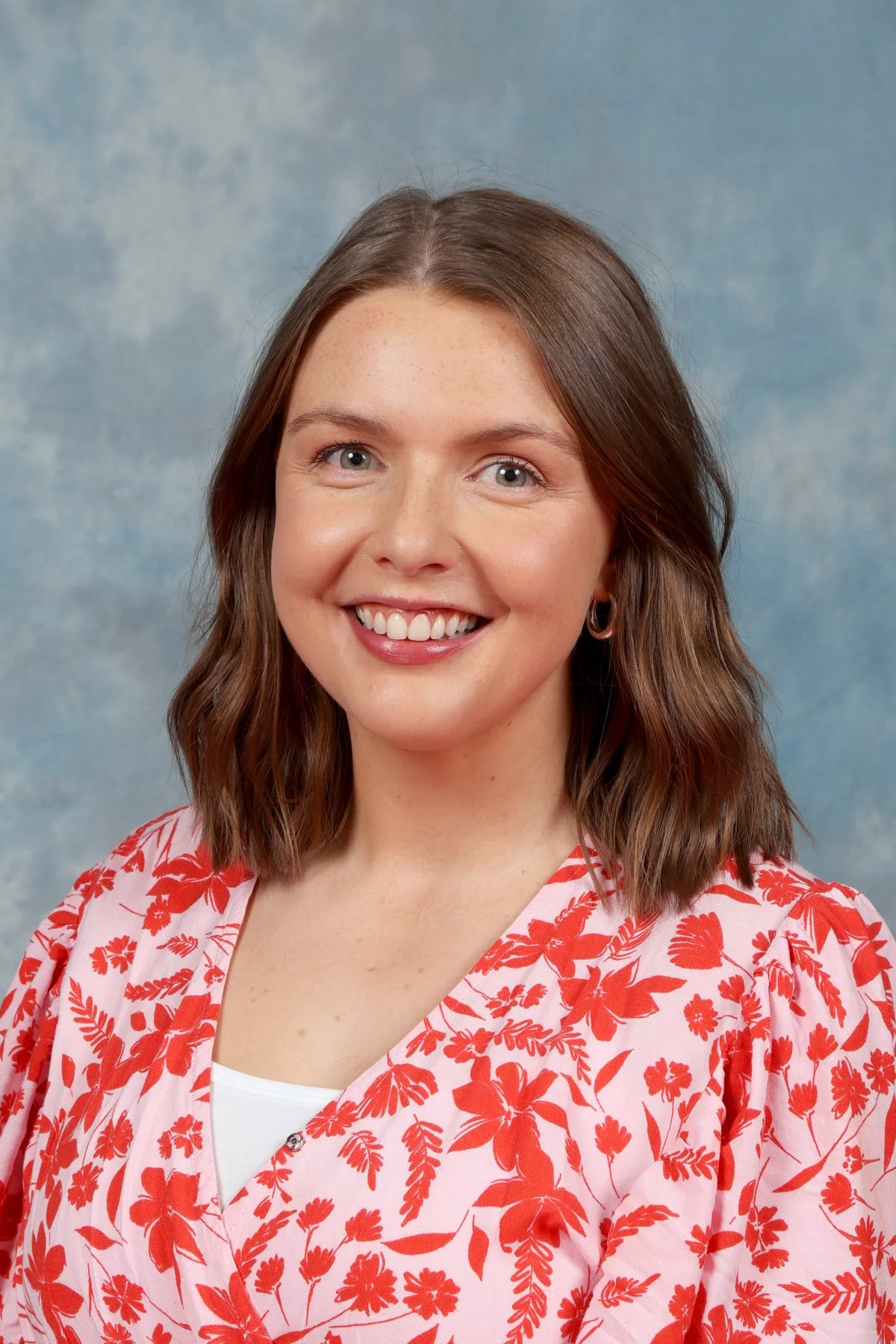 A woman with shoulder-length brown hair, blue eyes, and a wide smile, wearing a red and white floral blouse against a blue mottled background.