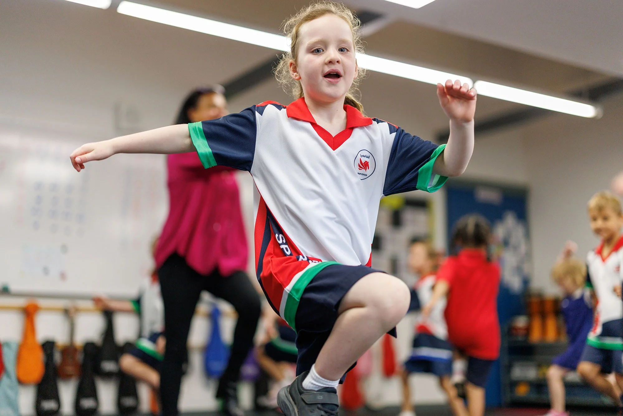 Young girl in sports uniform jumping in an indoor gym, with other children and a woman in the background.