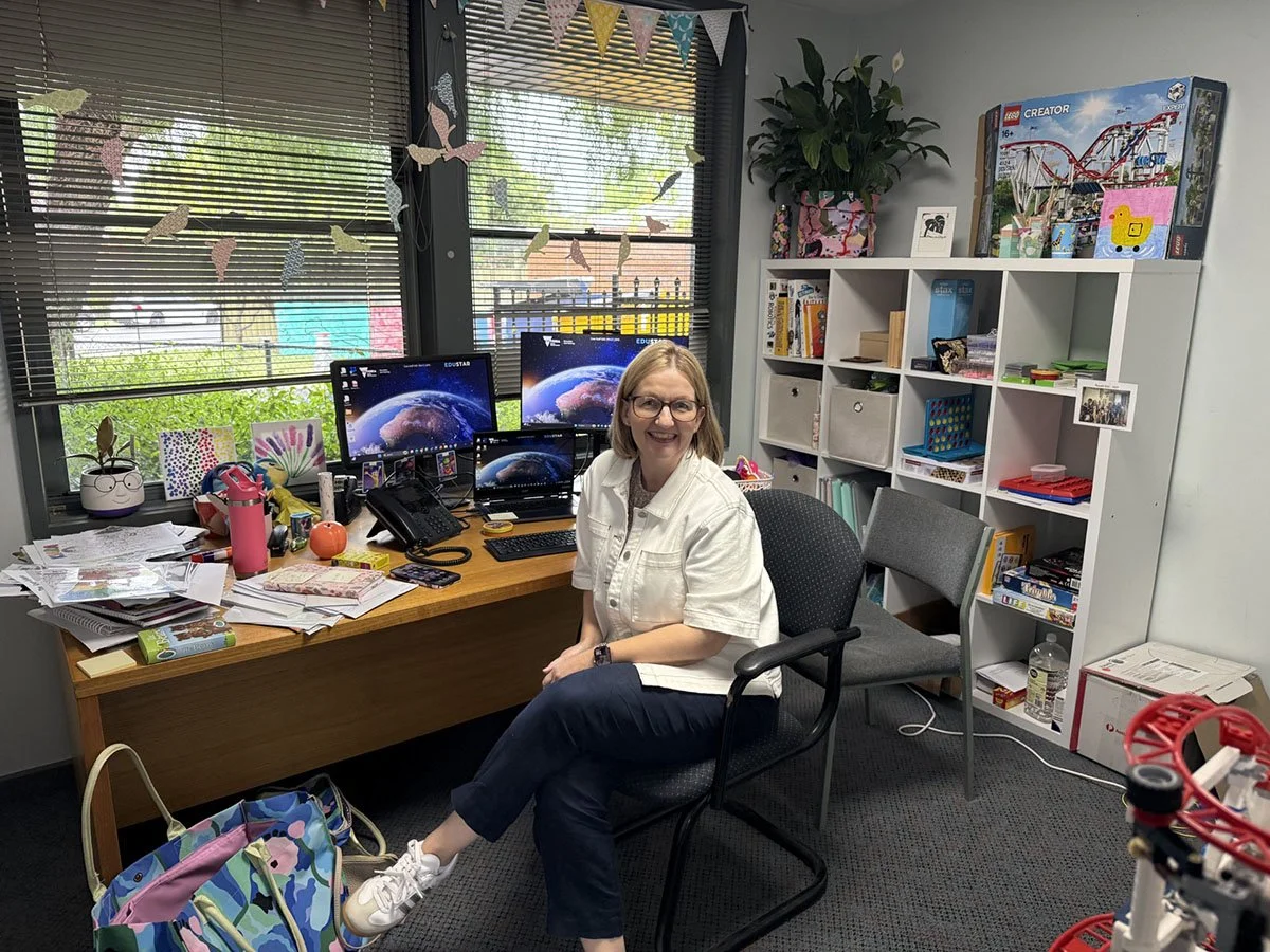 Woman sitting at a cluttered desk with two computer monitors in an office with window blinds, shelves, and colorful decorations.