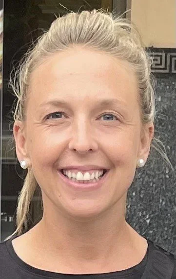 Close-up of a smiling woman with blonde hair tied back, wearing pearl earrings, standing outdoors.