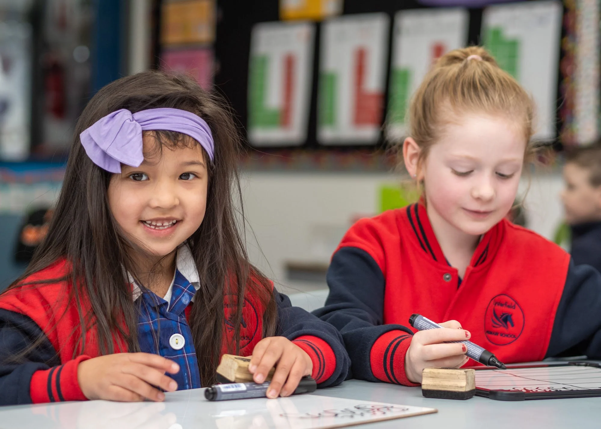 Two young school girls sitting at a table in classroom, smiling and engaged in writing activity with whiteboards and markers, colorful classroom background.