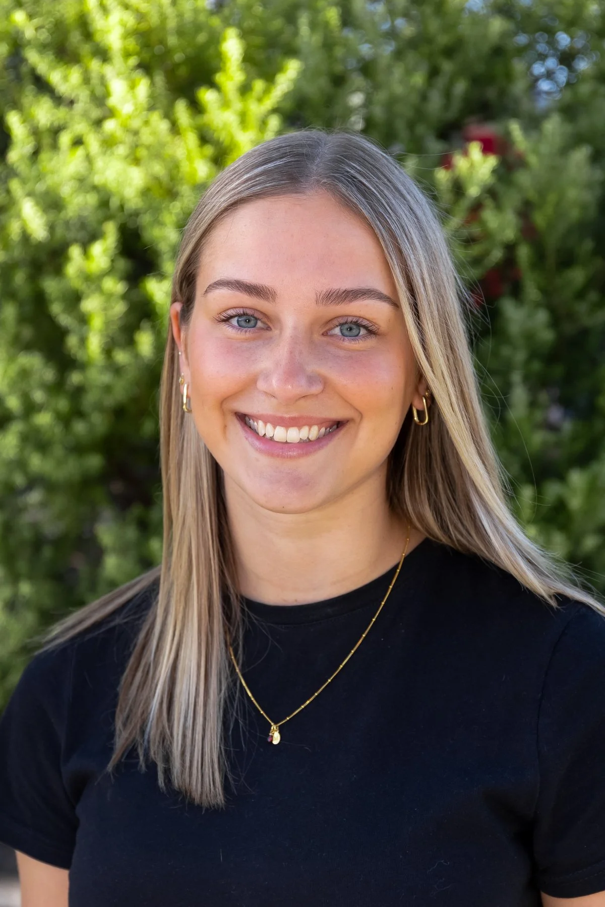 A young woman with blonde hair, blue eyes, and gold jewelry smiling outdoors with a background of green trees.