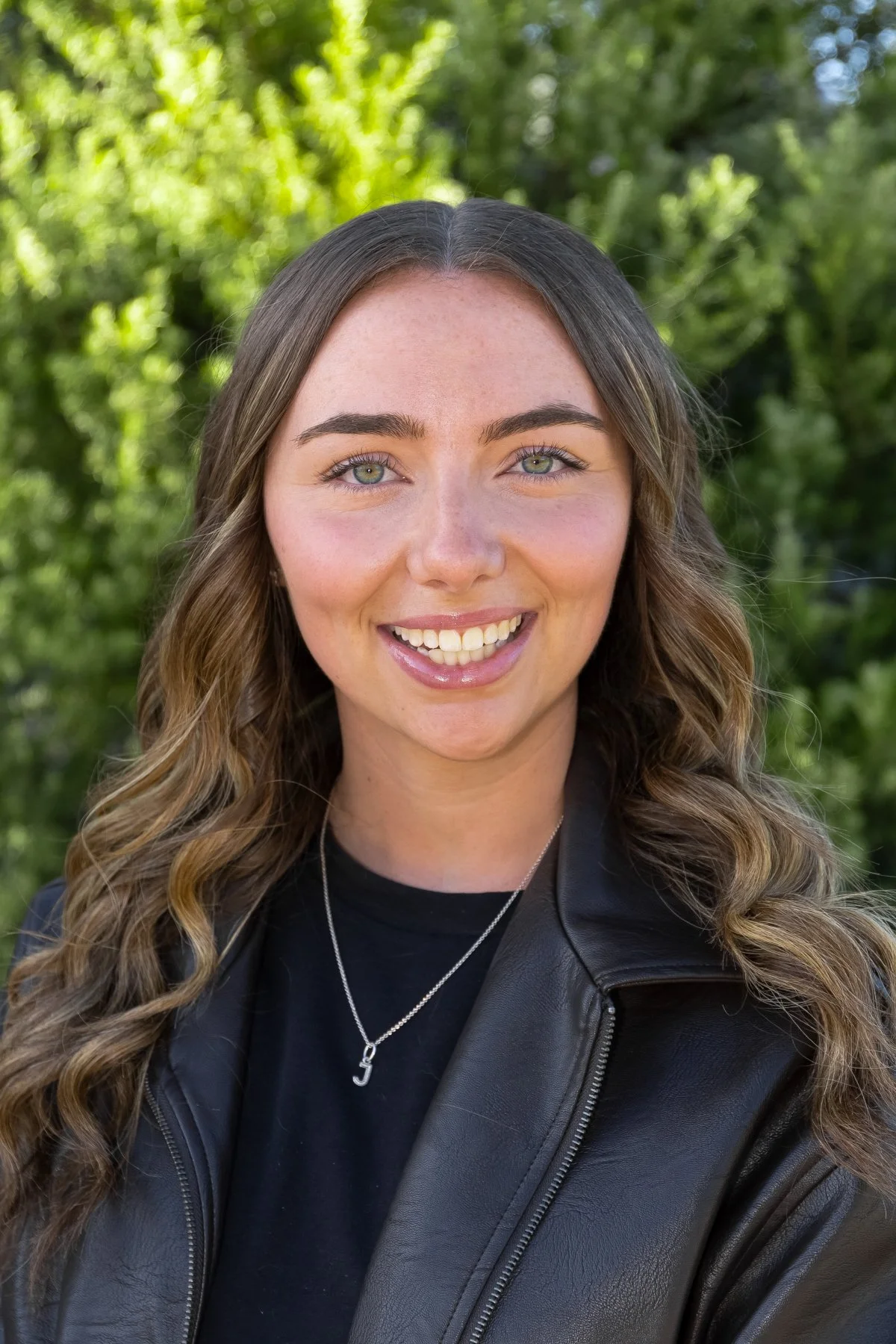 A young woman with long, wavy brown hair and blue eyes, smiling outdoors with greenery in the background, wearing a black leather jacket and a silver necklace with a letter J pendant.