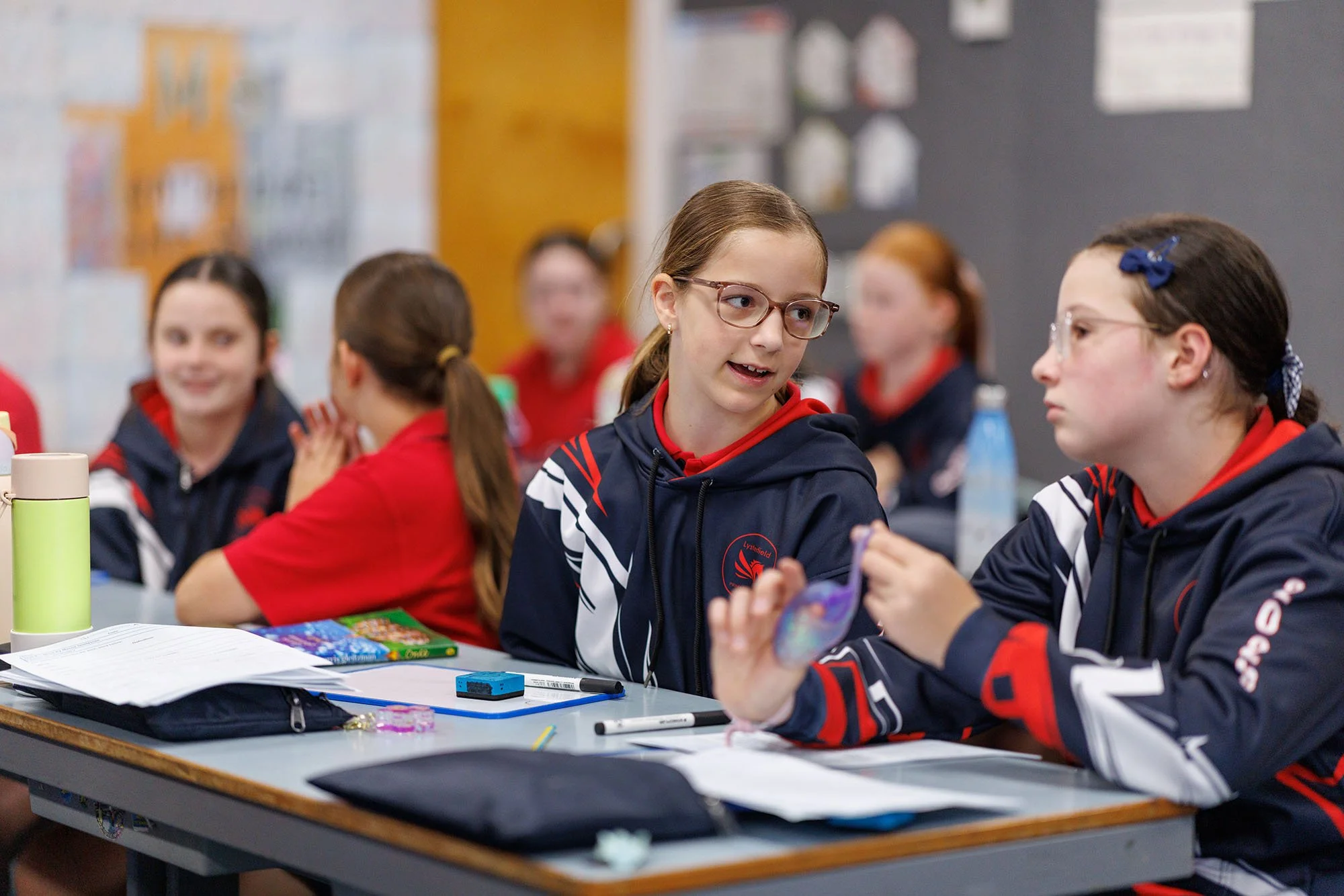 School classroom with students sitting at desks, engaging in conversation, some smiling, with educational posters on the walls.