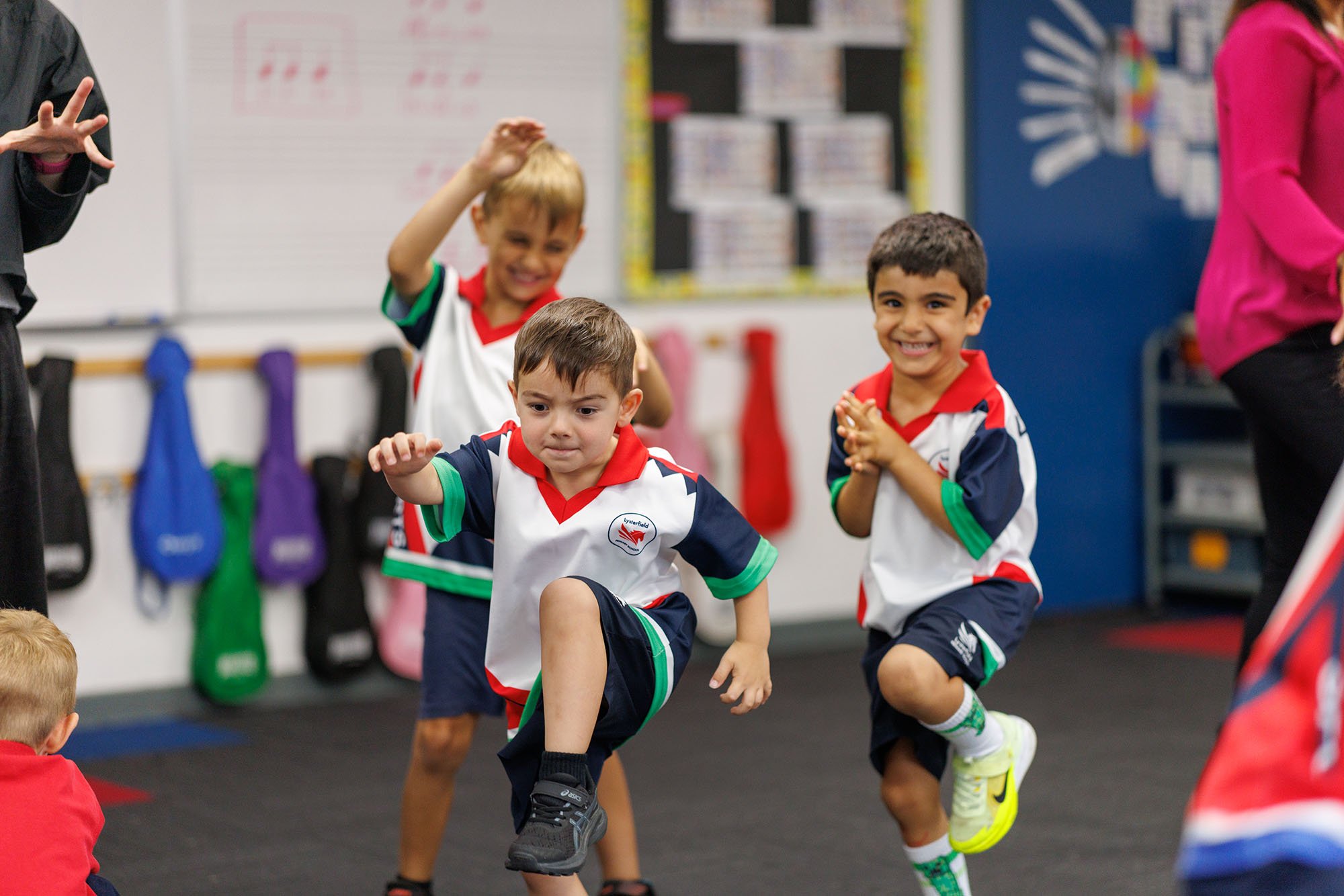 Young children participate in a physical activity or dance in a classroom, wearing matching sports uniforms, with some children smiling and one showing a serious expression.