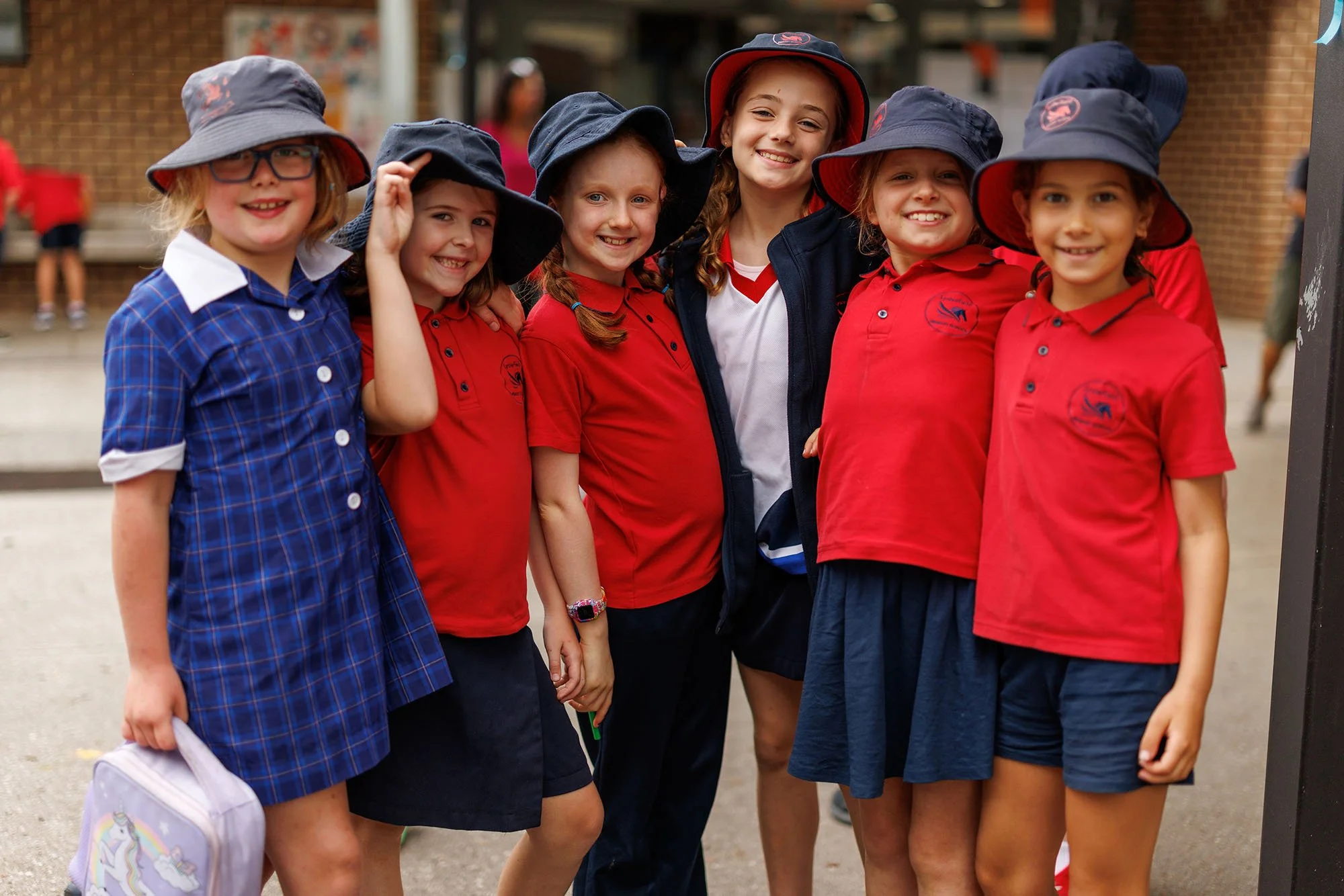 Group of six young girls in school uniform, smiling, wearing red shirts and navy hats, standing outdoors in a schoolyard.