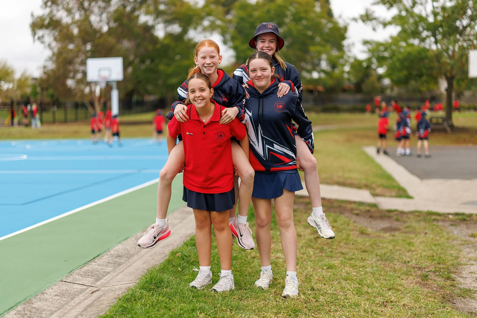 Group of young girls in sports uniforms, some piggybacking others, smiling outdoors on a grassy area near a blue court, with other children in the background.