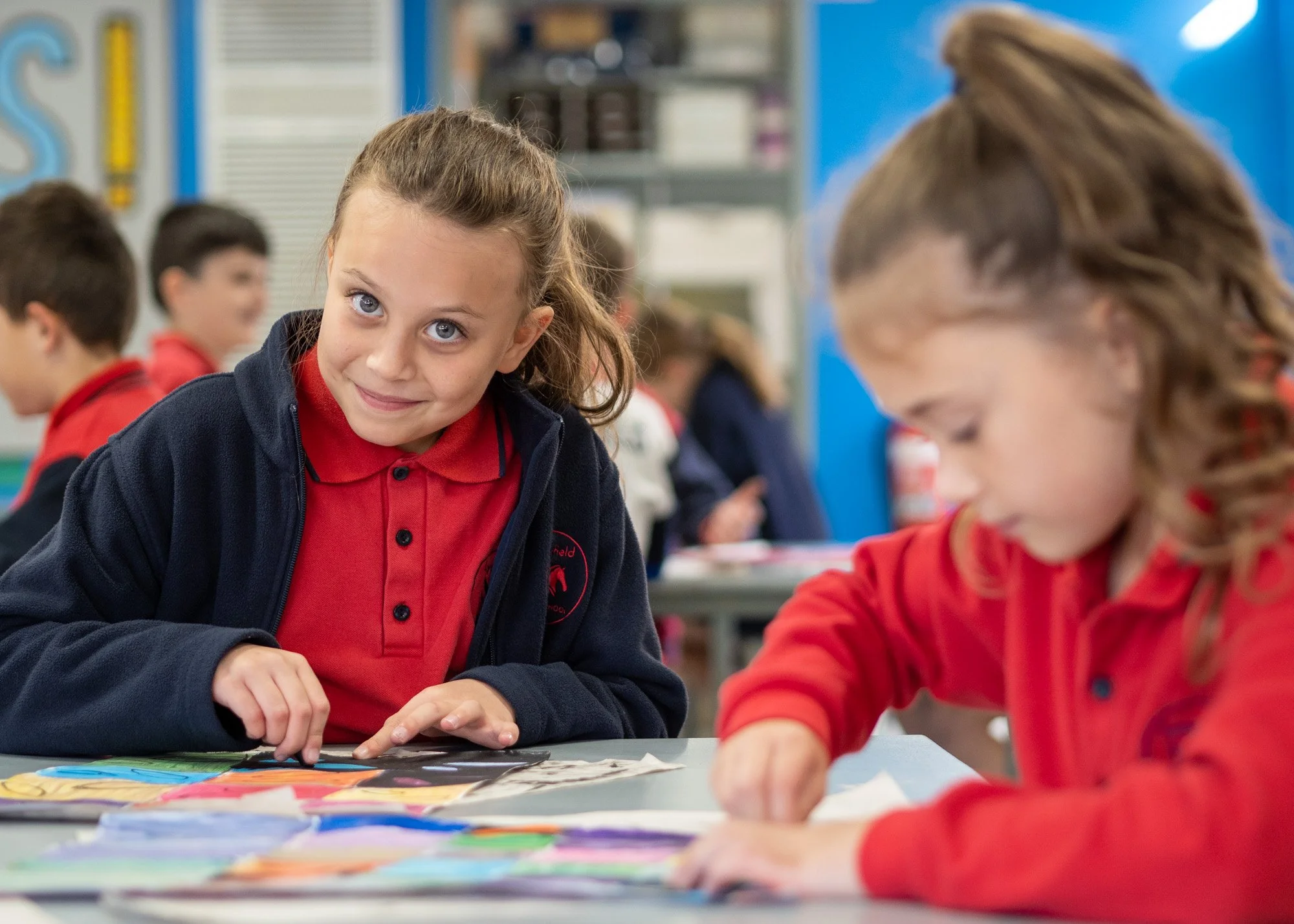 A young girl with brown hair pulled back, smiling and wearing a red polo shirt and a navy jacket, looking at the camera while sitting at a table with colorful papers or artwork in a classroom. Other children are in the background, engaged in activities.