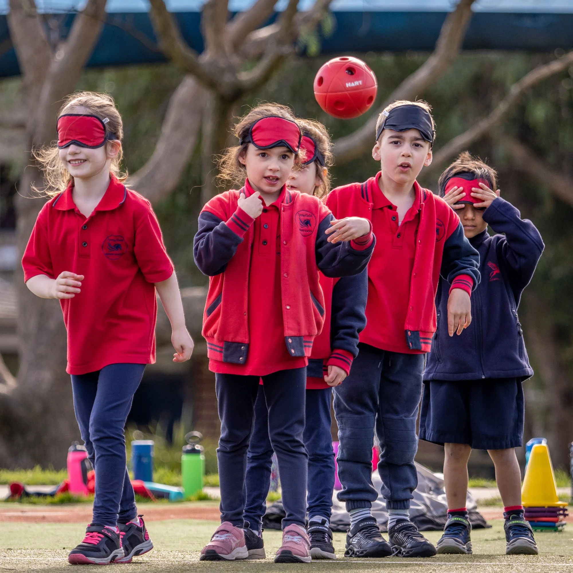 Children participate in an outdoor activity, with four kids wearing red and navy blue school uniforms, some blindfolded, alerting about a red ball in the air. In the background, trees and outdoor gear are visible.