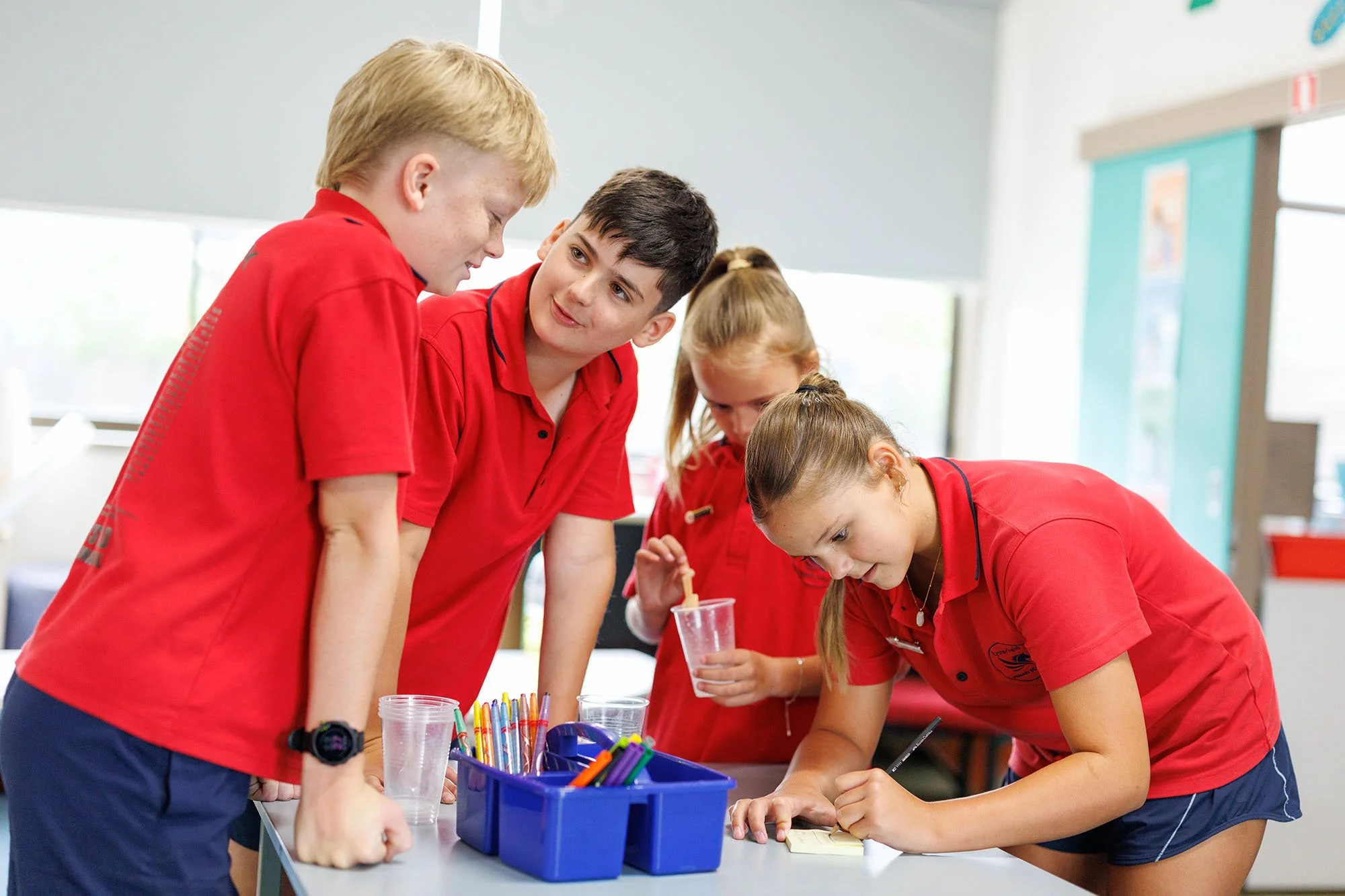 Four children in red uniforms gather around a table, with a girl writing on a notepad while the others observe, in a classroom setting.