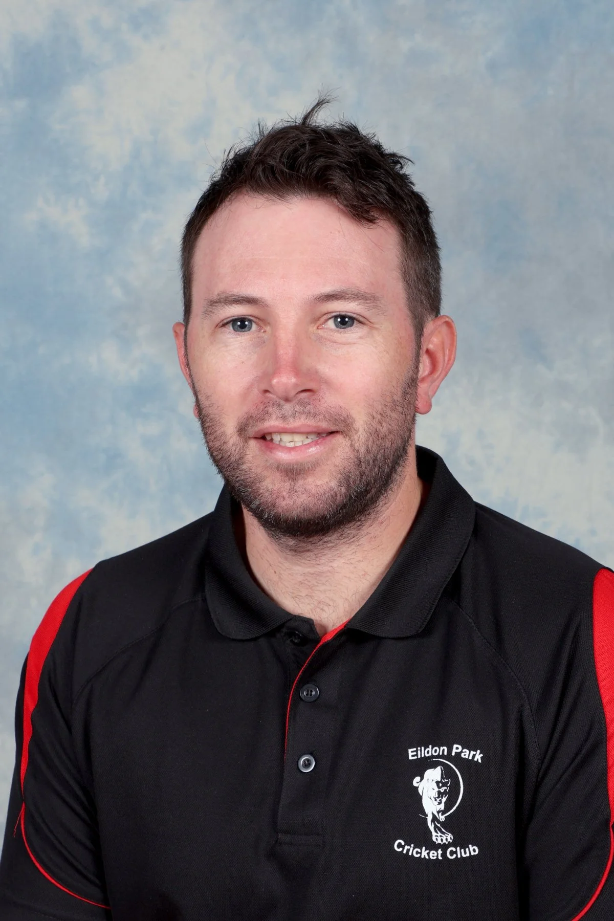 A man with short dark hair and a beard wearing a black and red polo shirt with the logo of Eildon Park Cricket Club.