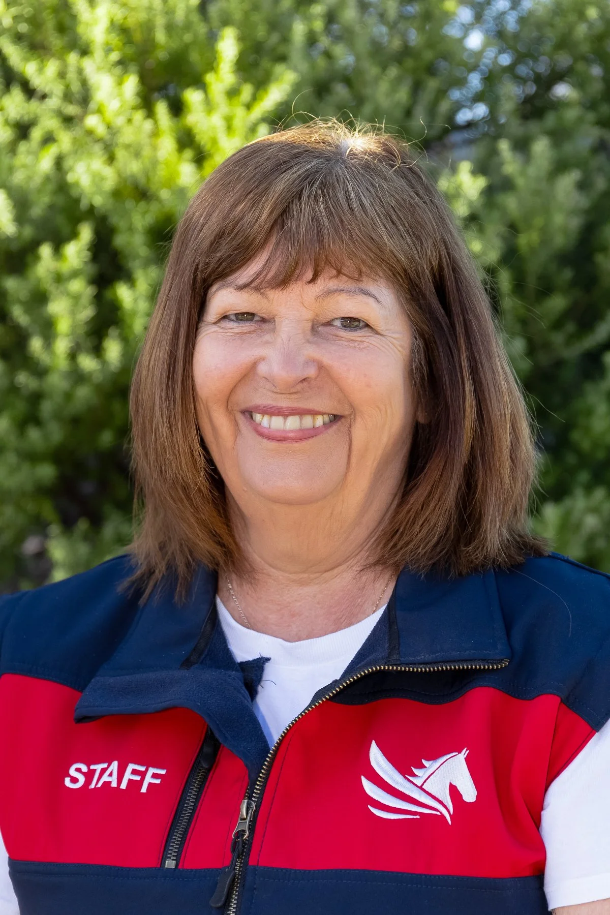 A smiling woman with shoulder-length brown hair standing outdoors with green trees in the background. She is wearing a navy and red jacket with a white horse logo and the word 'STAFF' on it.