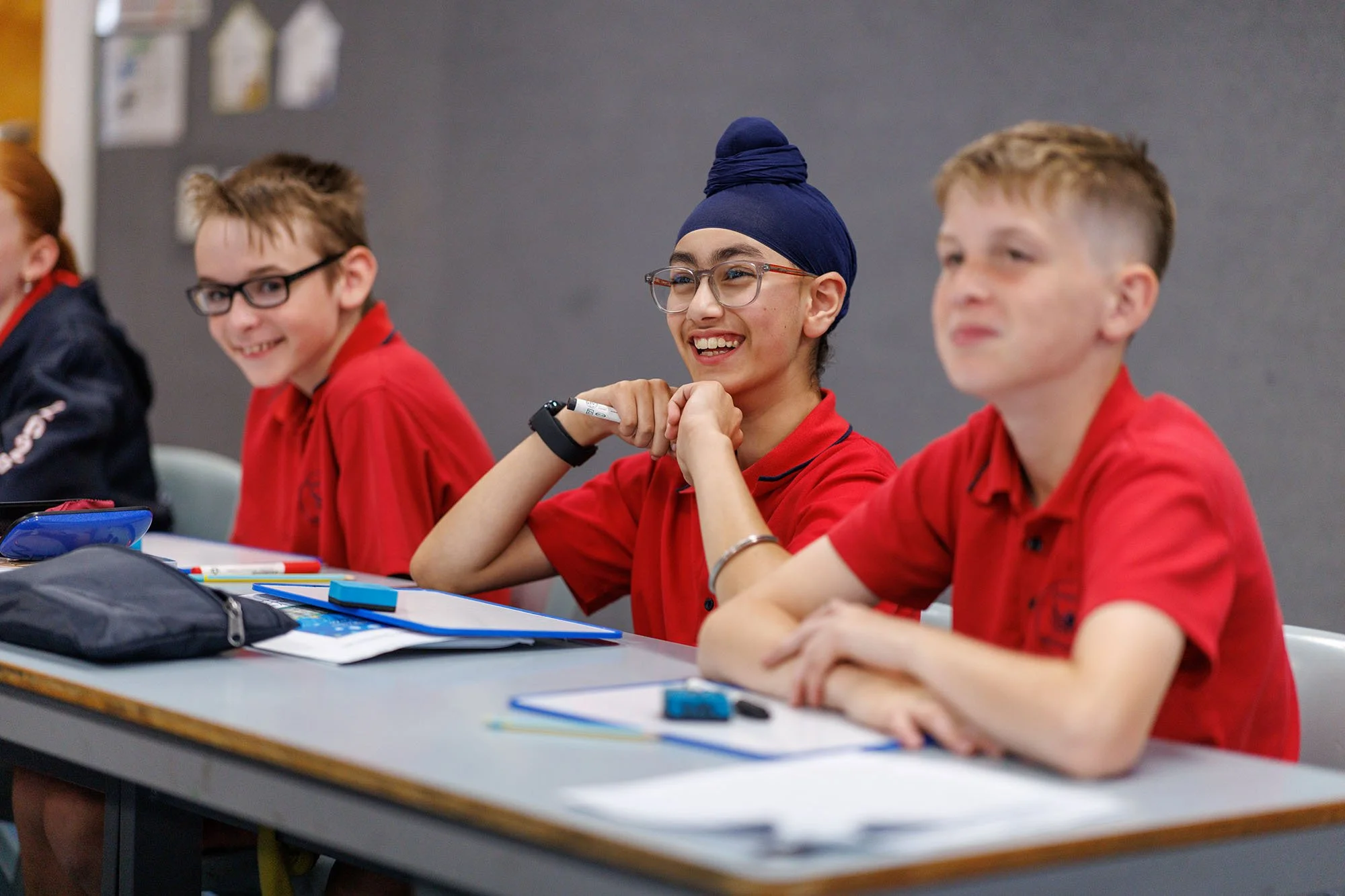 Students sitting at desks in a classroom, smiling, dressed in red shirts, with school supplies on the desk.