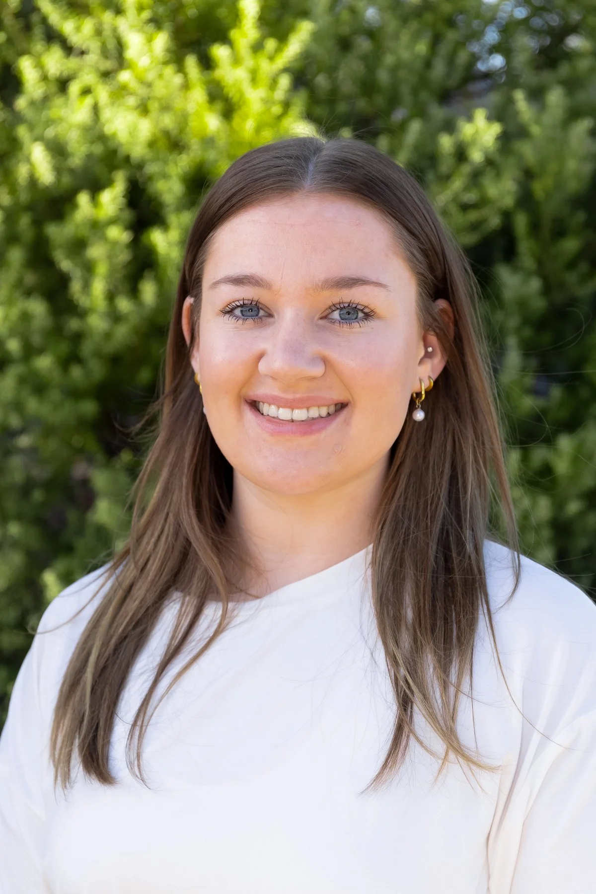 A young woman with long brown hair and blue eyes smiling outside with green trees in the background, wearing a white top and pearl earrings.