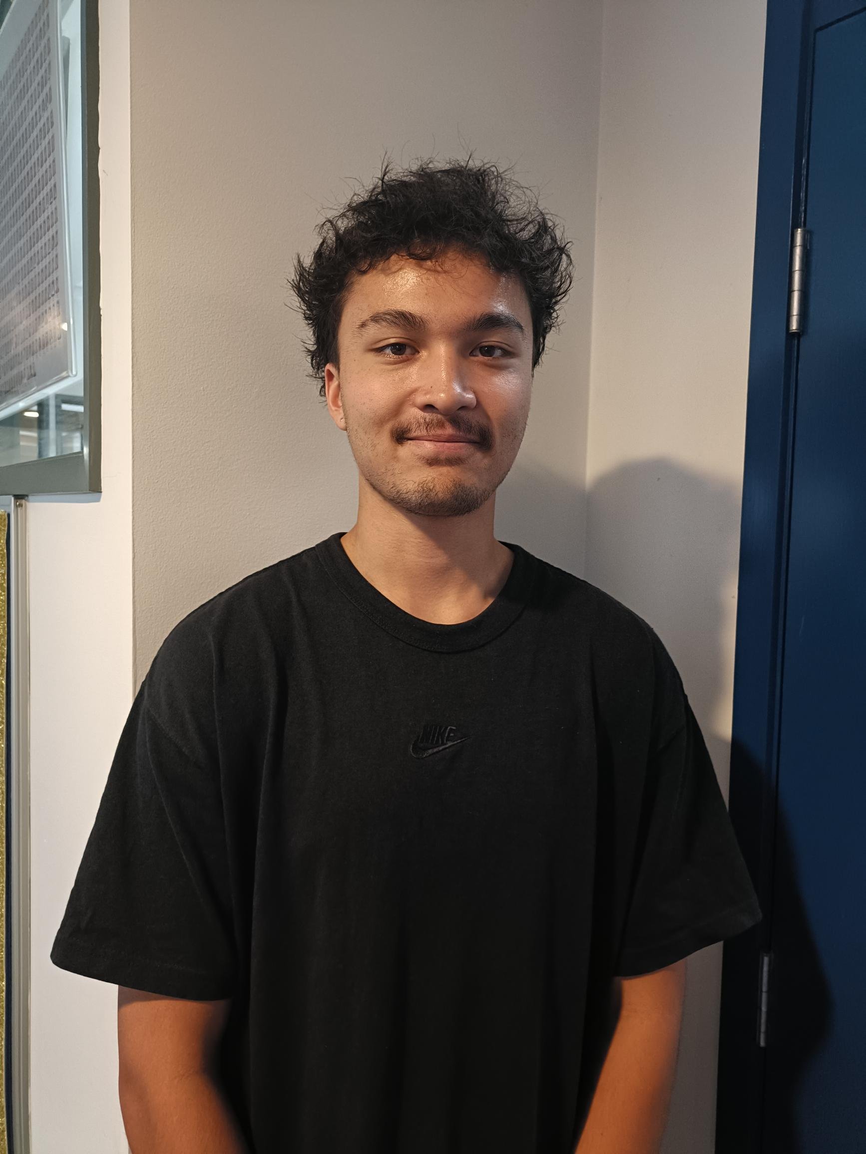 A young man with curly dark hair, light facial hair, and wearing a black Nike t-shirt, standing indoors against a beige wall.