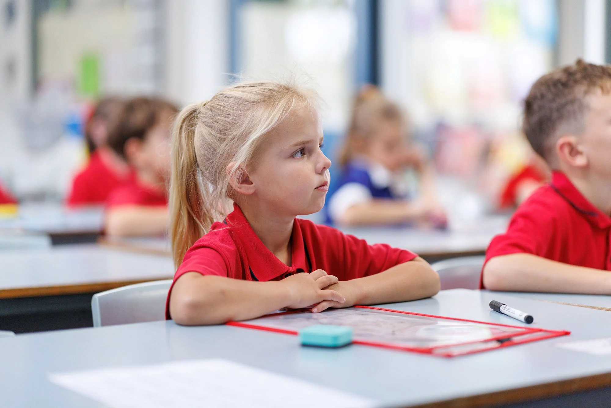 A young girl with blonde hair sitting at a desk in a classroom, attentively listening, surrounded by other children.