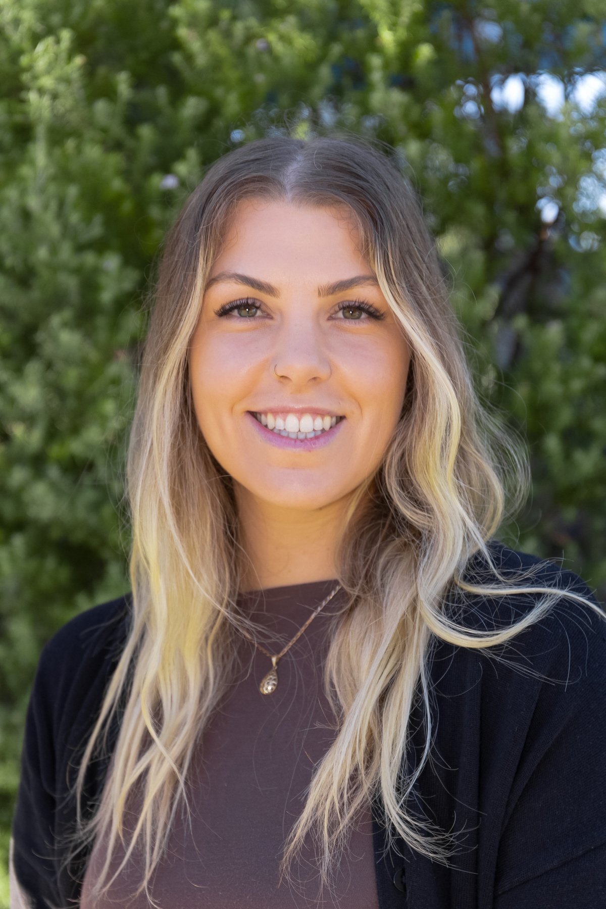 A young woman with long, wavy blonde hair, smiling, outdoors with green trees in the background.