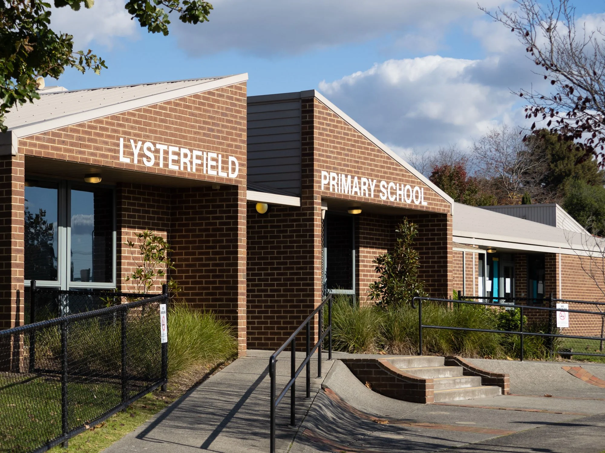 Exterior of Lysterfield Primary School building with brick walls, set against a partly cloudy sky and trees.