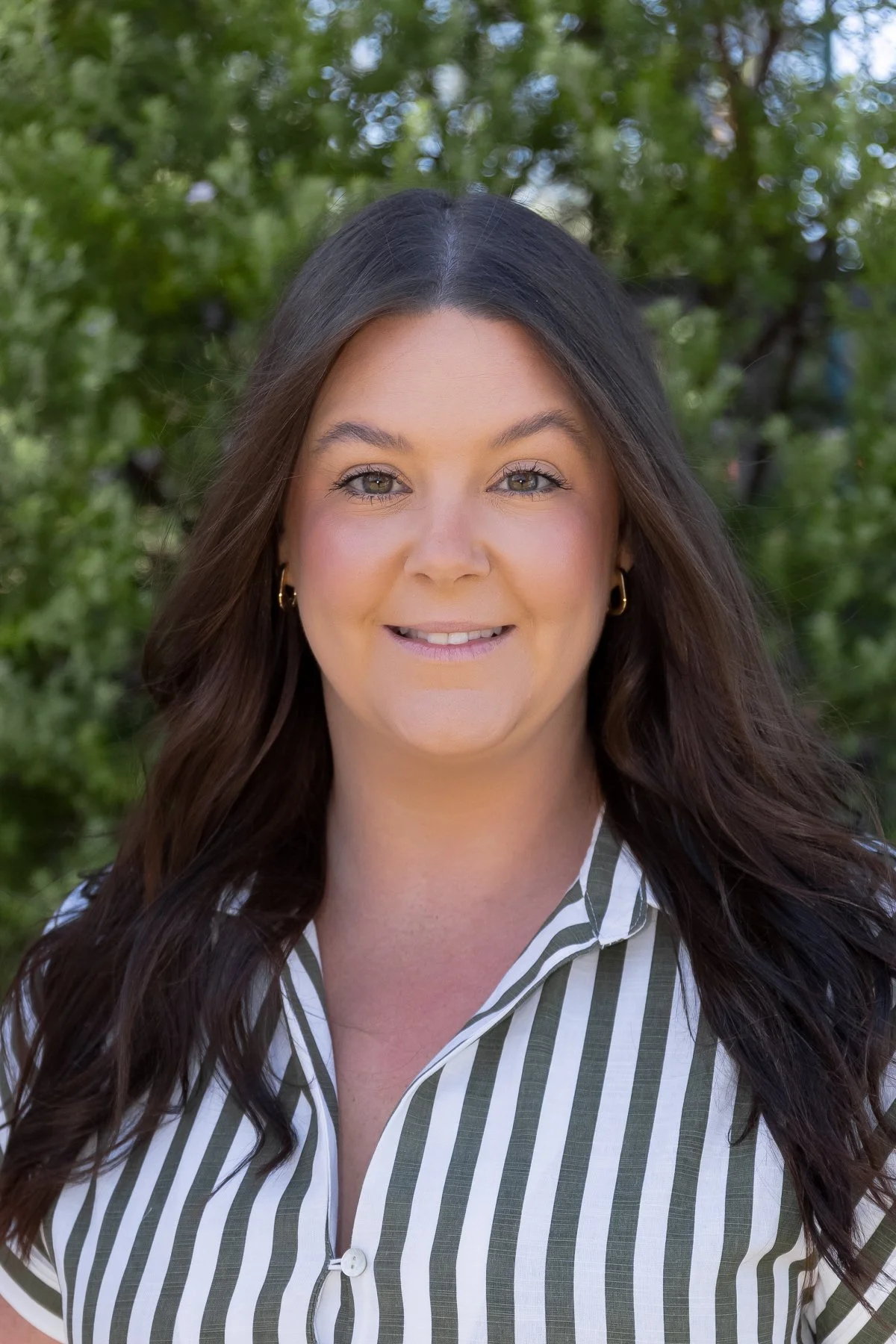 A woman with long brunette hair, wearing a white and green striped shirt, smiling outdoors with green trees in the background.
