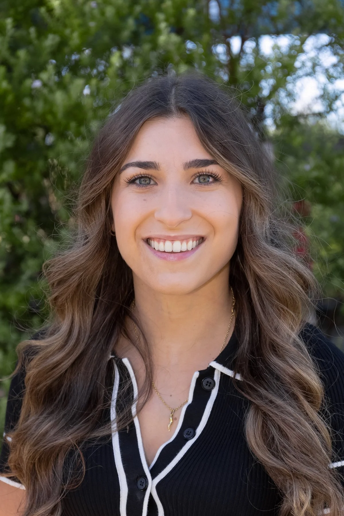 A young woman with long wavy brown hair, blue eyes, and a bright smile standing outdoors with green trees in the background.