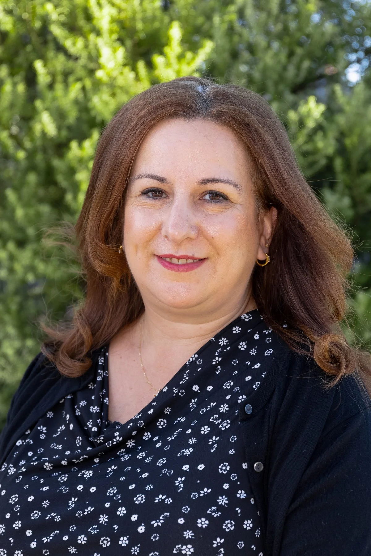 A woman with shoulder-length reddish-brown hair and light skin, smiling at the camera, standing outdoors with green trees in the background.