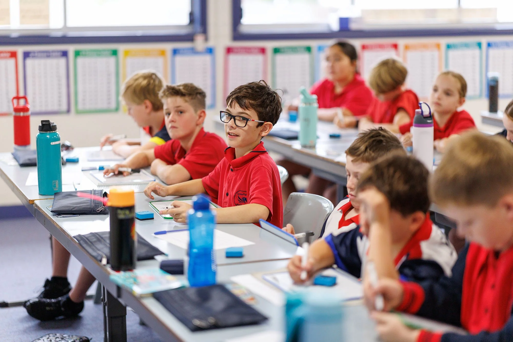 Students in a classroom sitting at desks wearing red uniforms with water bottles and school supplies, engaging in lesson activities.