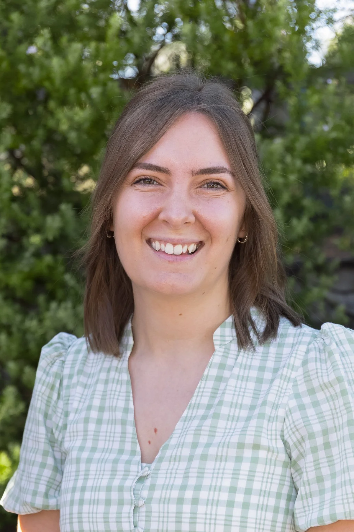 A young woman with shoulder-length brown hair smiling outdoors, wearing a light green and white checkered top with a zip detail, with green trees in the background.