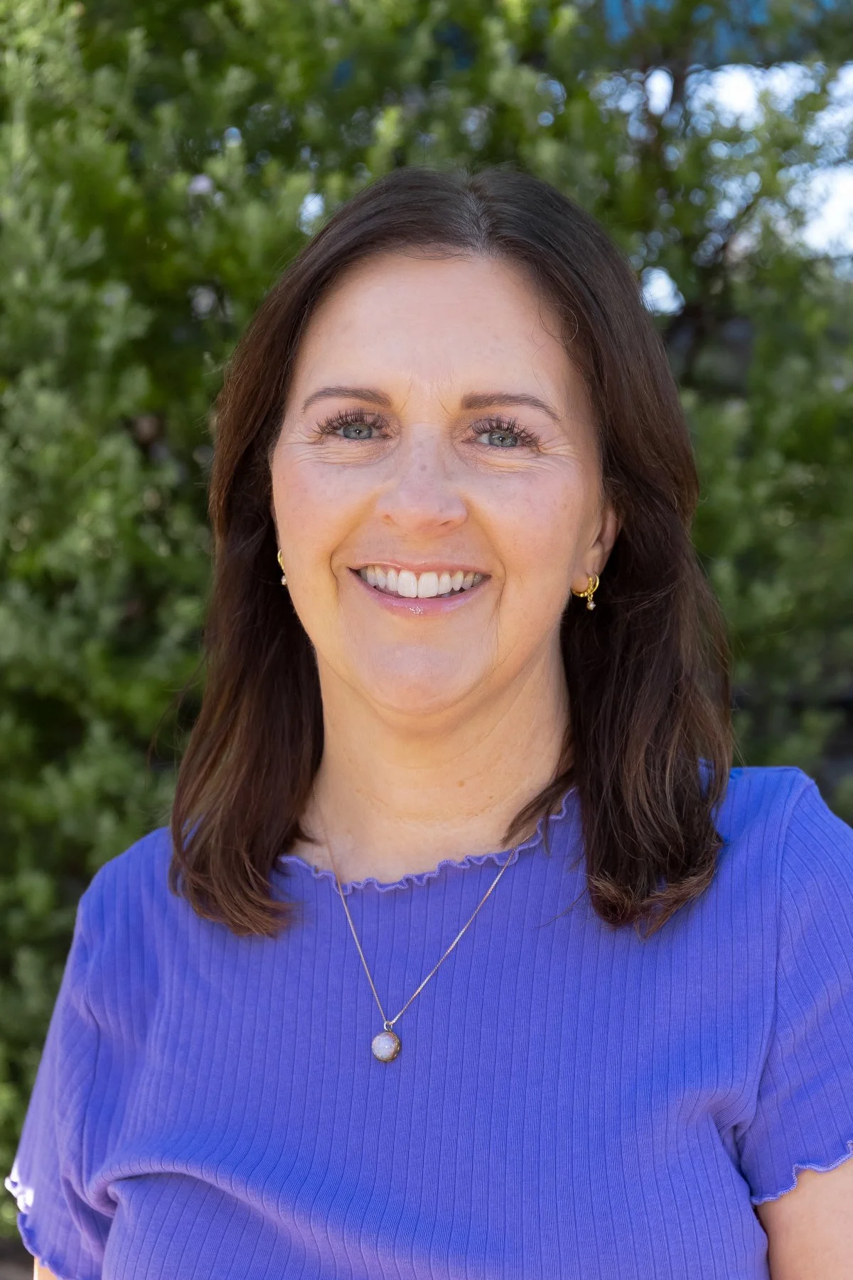 A smiling woman with shoulder-length brown hair, wearing a blue top, gold earrings, and a necklace with a circular pendant, standing outdoors with green trees in the background.