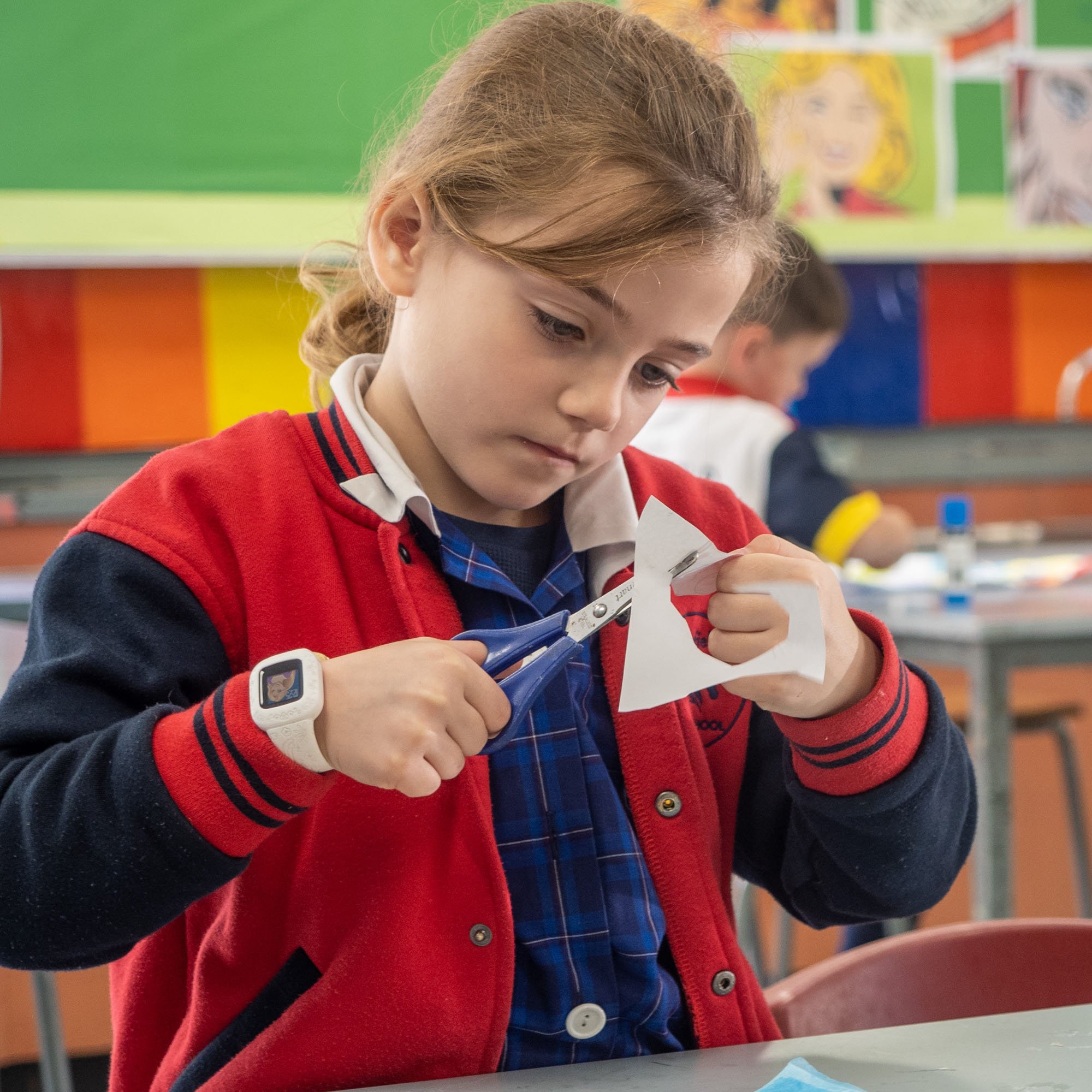 A young girl in a school uniform and red jacket cutting paper with scissors in a classroom.