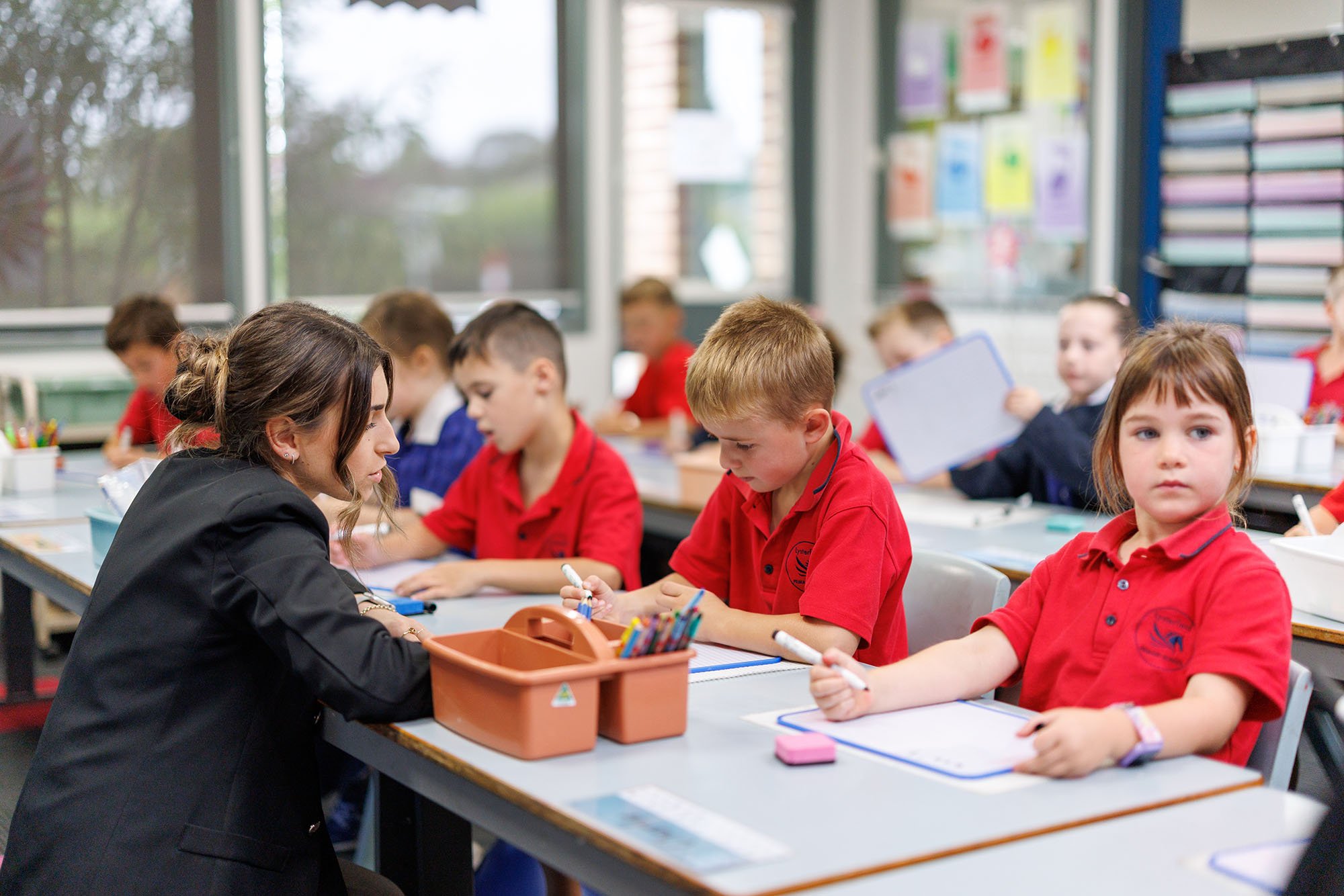 Teacher assisting students in a classroom with red uniforms, sitting at desks with papers and writing supplies.