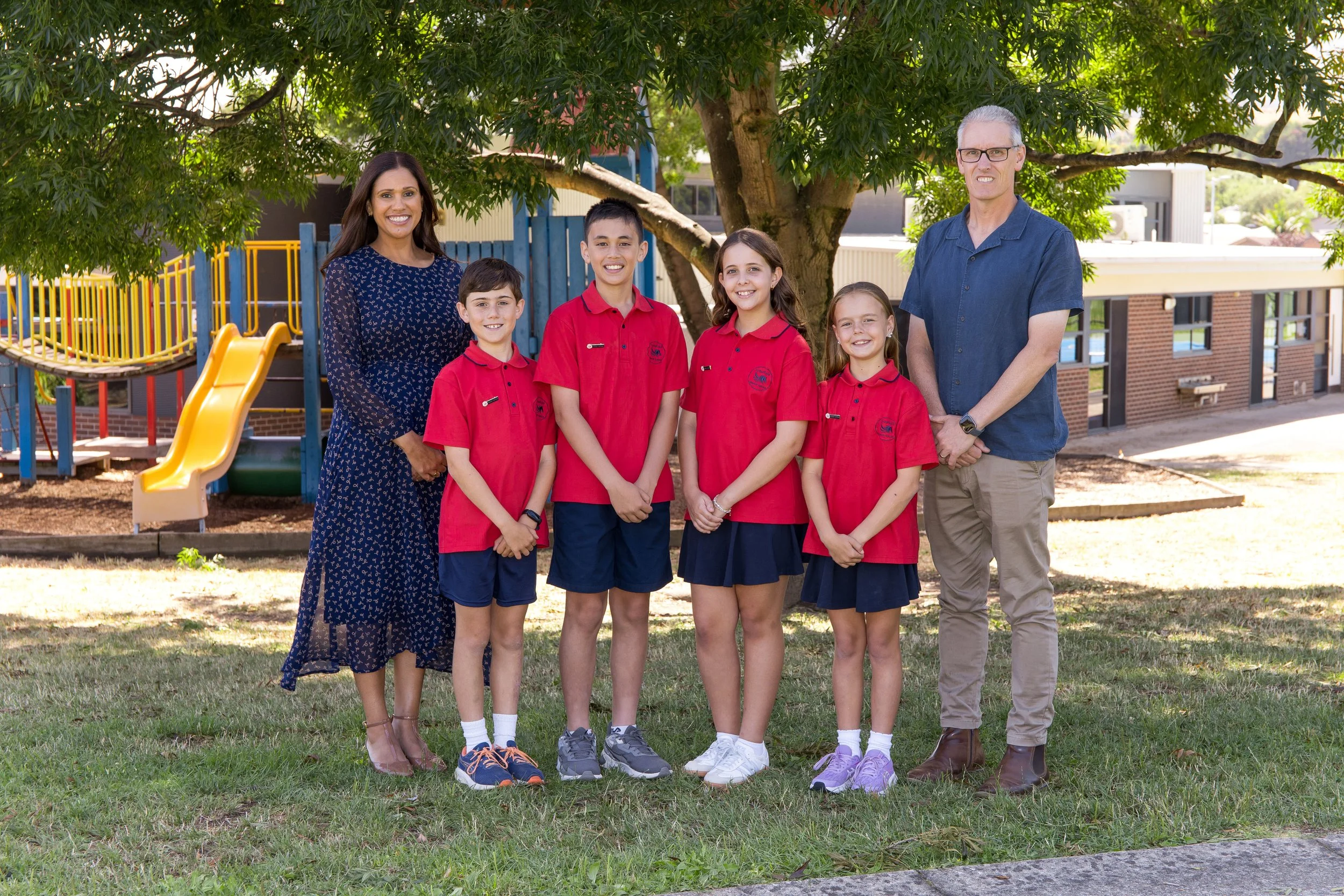 A group of two adults and four children stands outdoors in front of a large tree, a playground with a yellow slide behind them. The children are wearing red school uniforms, and the adults are dressed casually. All are smiling at the camera.