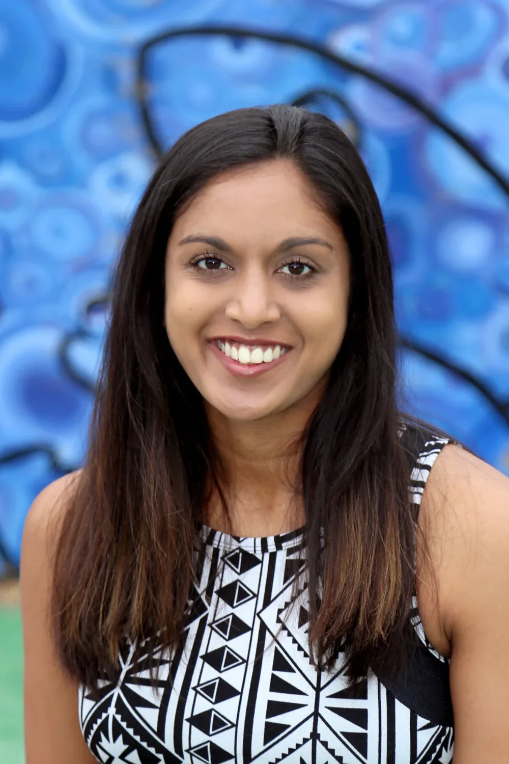 A young woman with long dark hair smiling in front of a blue graffiti wall.