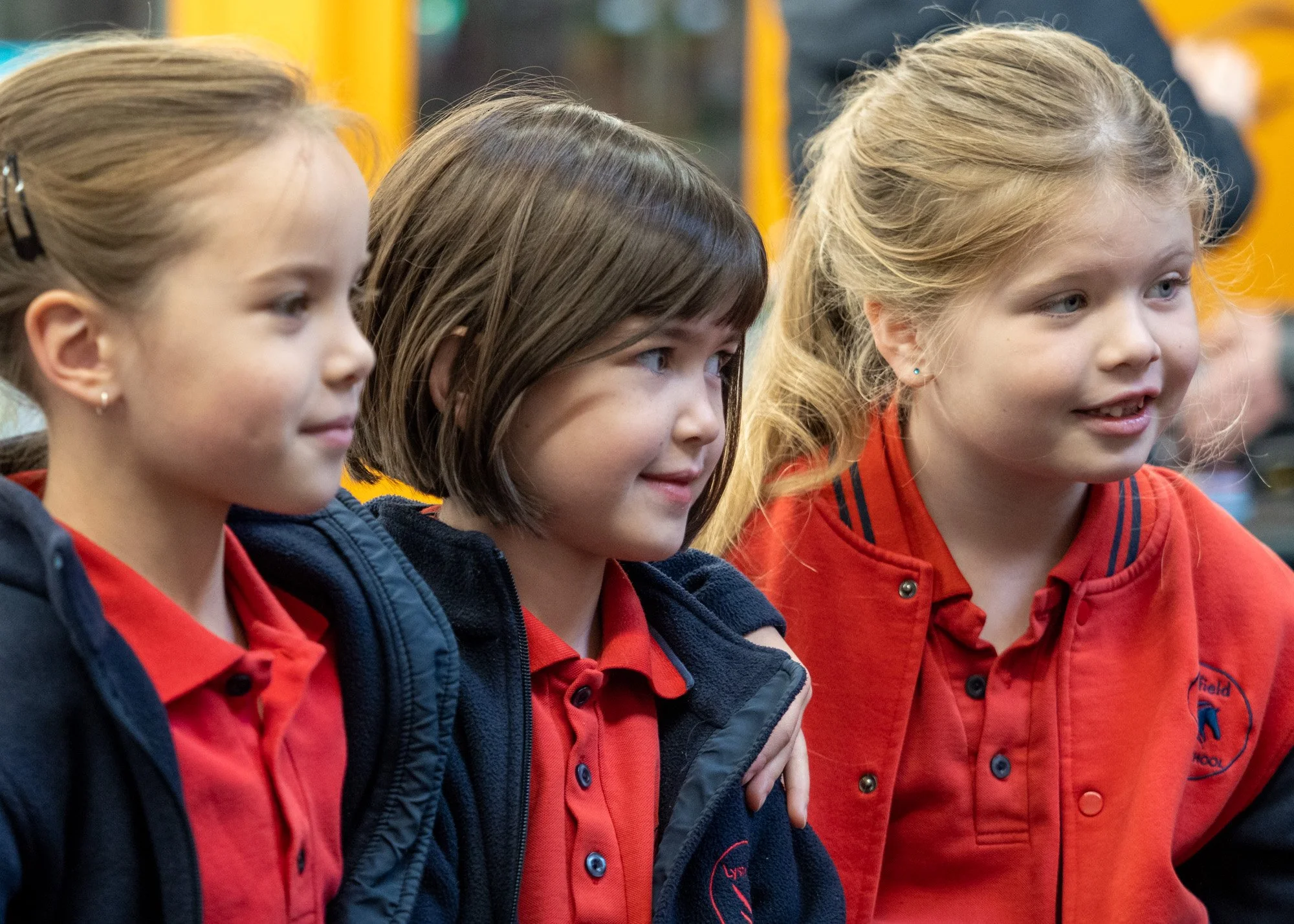 Three young girls in red school uniforms standing together, smiling, inside a school hallway.