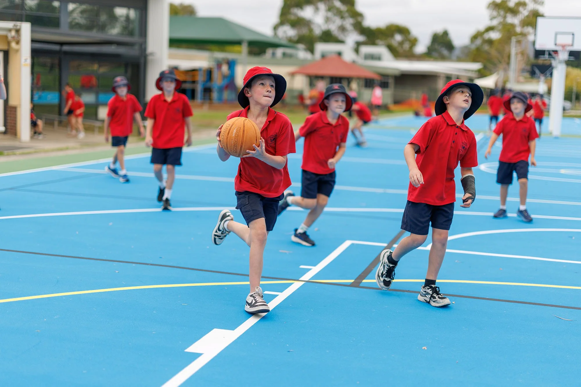 Children in red shirts and hats playing basketball on an outdoor court.