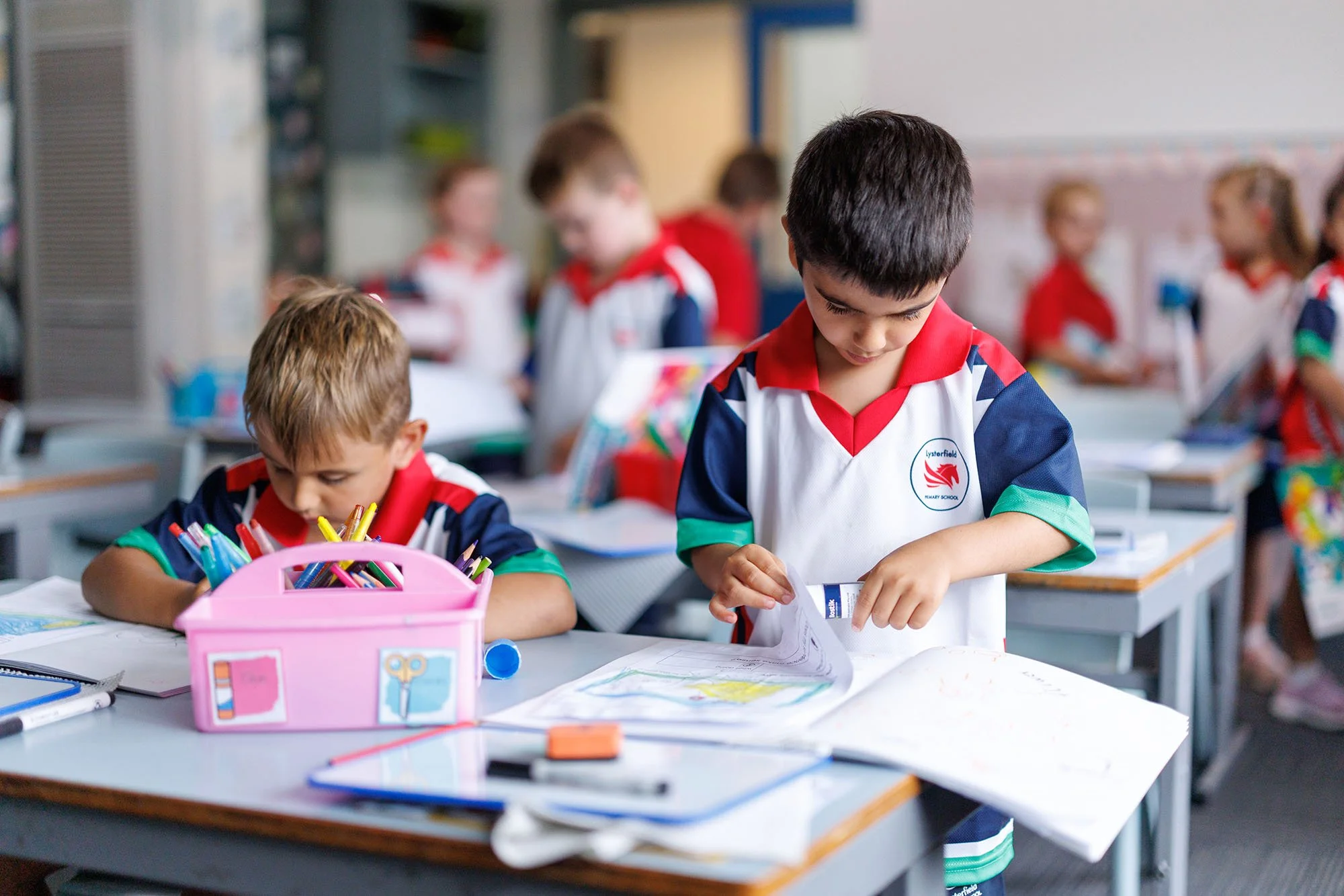 Two young boys in a classroom wearing school uniforms, one looking at a paper and the other writing, with school supplies on the desks and other children in the background.