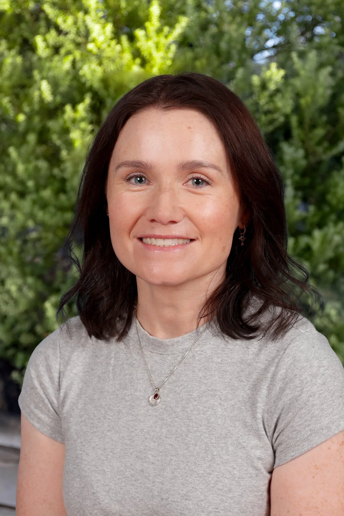 A woman with shoulder-length dark hair smiling outdoors with green trees in the background.