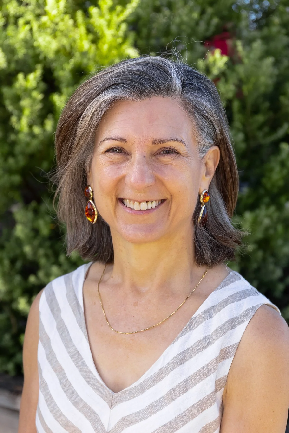 A smiling woman with shoulder-length gray hair wearing earrings, a necklace, and a sleeveless striped top, standing outdoors with greenery in the background.