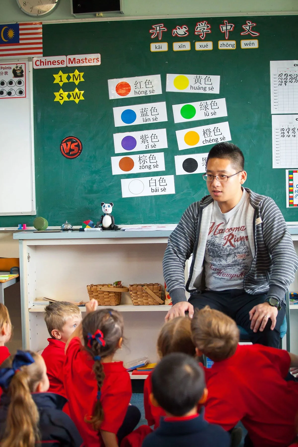 A classroom with children in red uniforms sitting on the floor, facing a teacher. The blackboard behind the teacher has colorful Chinese characters and color swatches with names and pronunciation. The classroom has educational posters, and a small panda figurine and other items are on the desk.