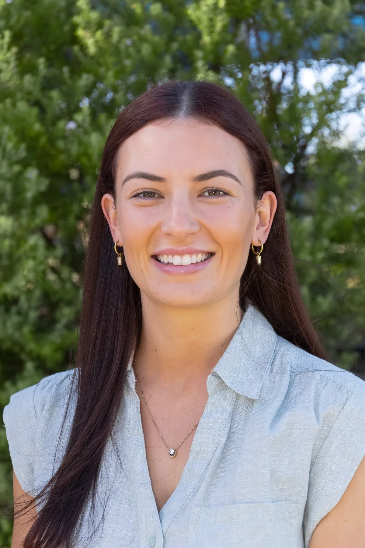 A woman smiling outdoors with green trees in the background.