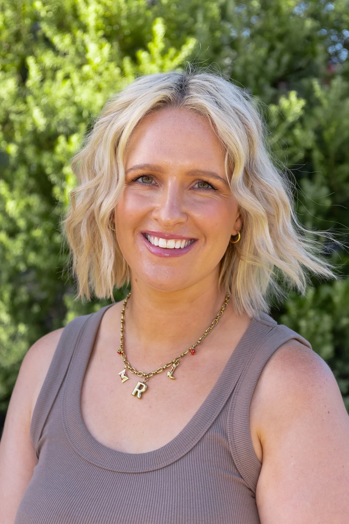 A woman with blonde, wavy hair and green eyes, smiling outdoors with trees in the background, wearing a sleeveless gray top and gold jewelry including a necklace with letters and small charms.