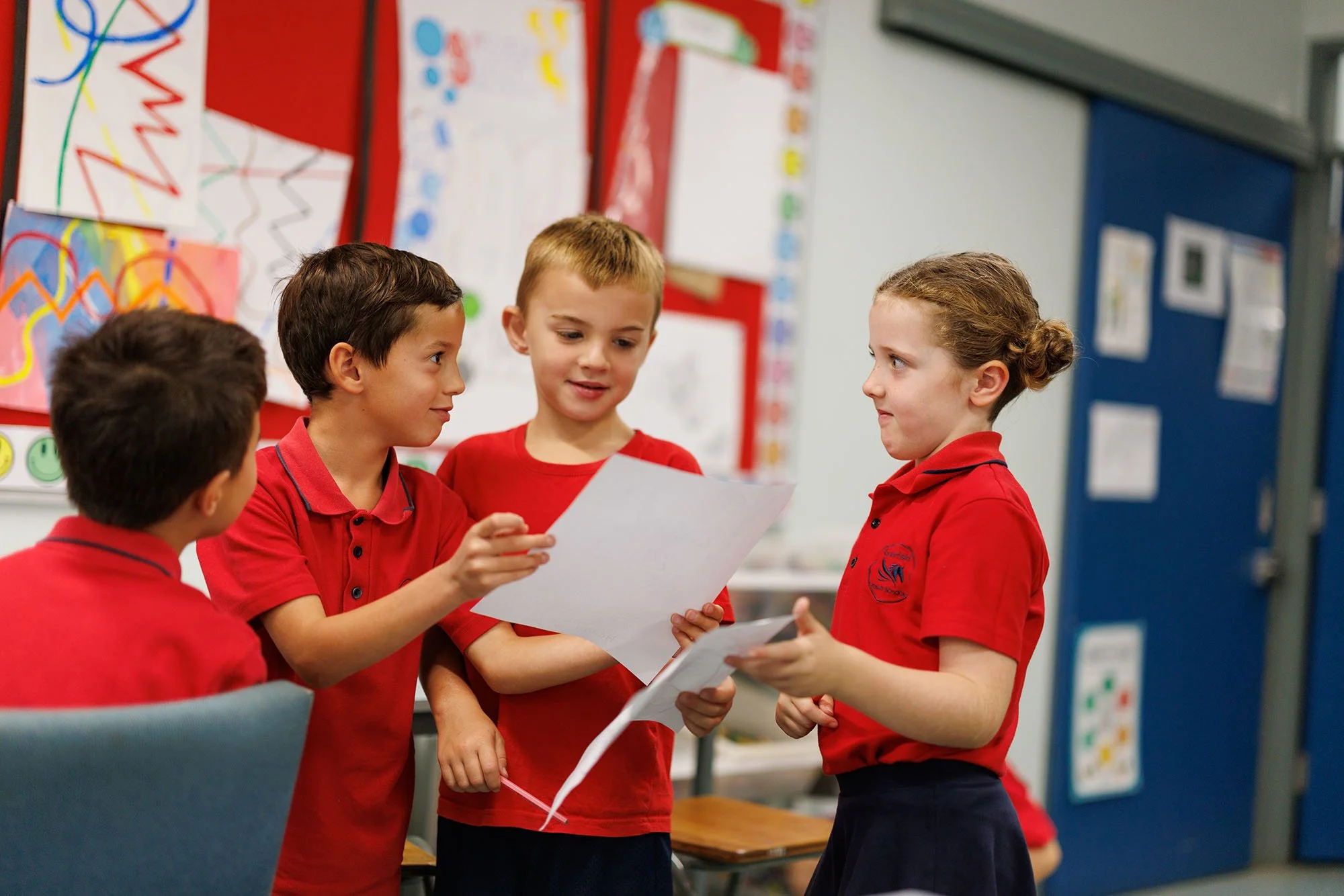 Four children wearing red shirts are having a discussion in a classroom, with colorful artwork on the walls.
