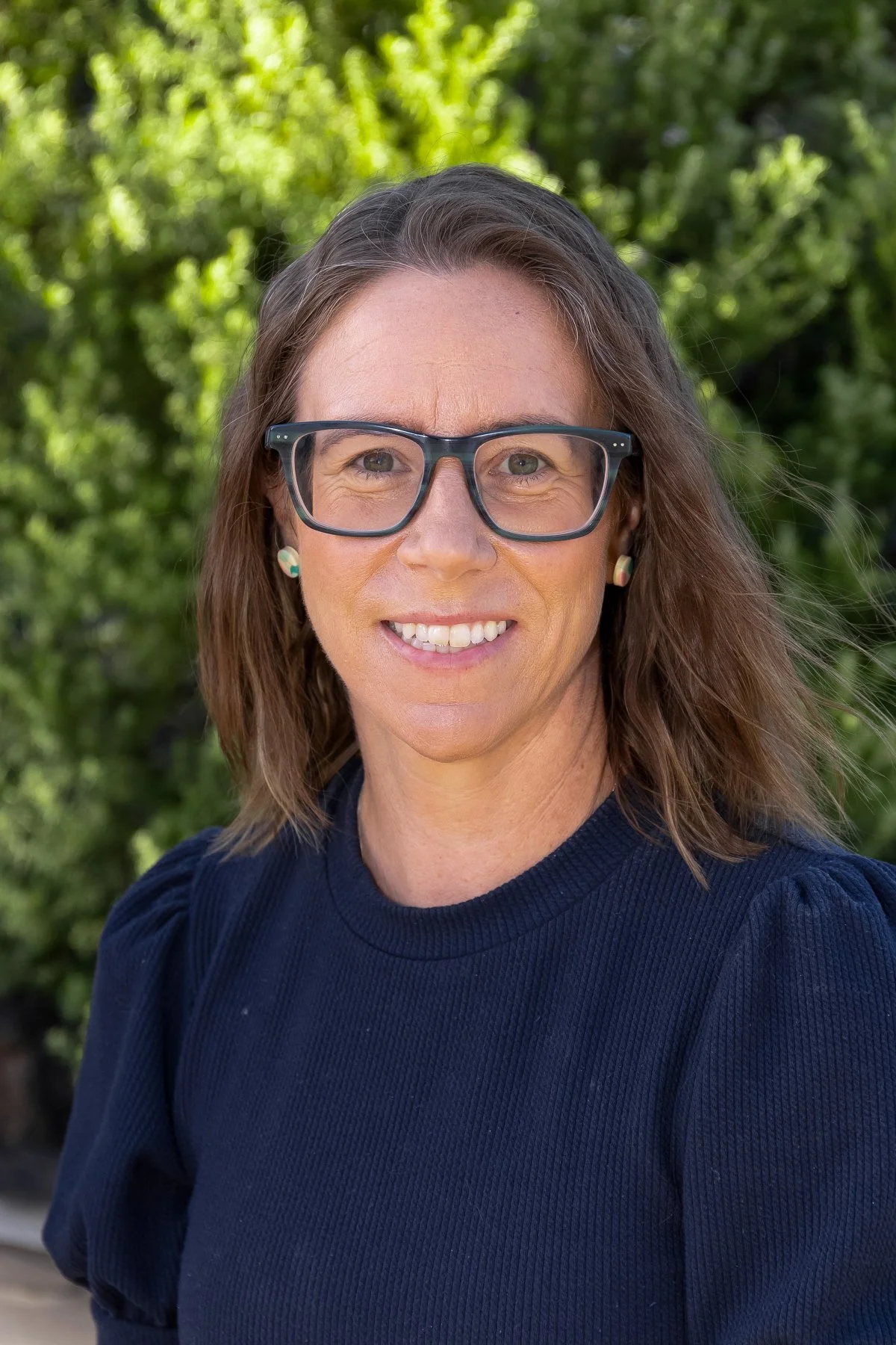 A woman with shoulder-length brown hair, wearing glasses and earrings, smiling outdoors with green trees in the background.