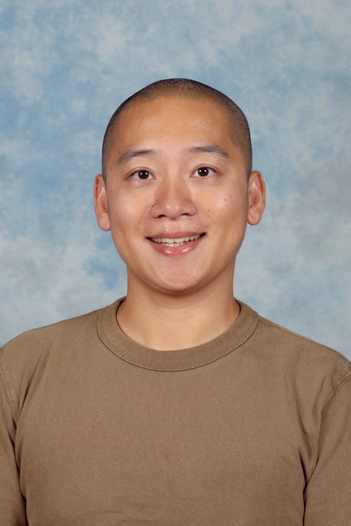 A young man with short hair smiling in front of a blue cloudy background.