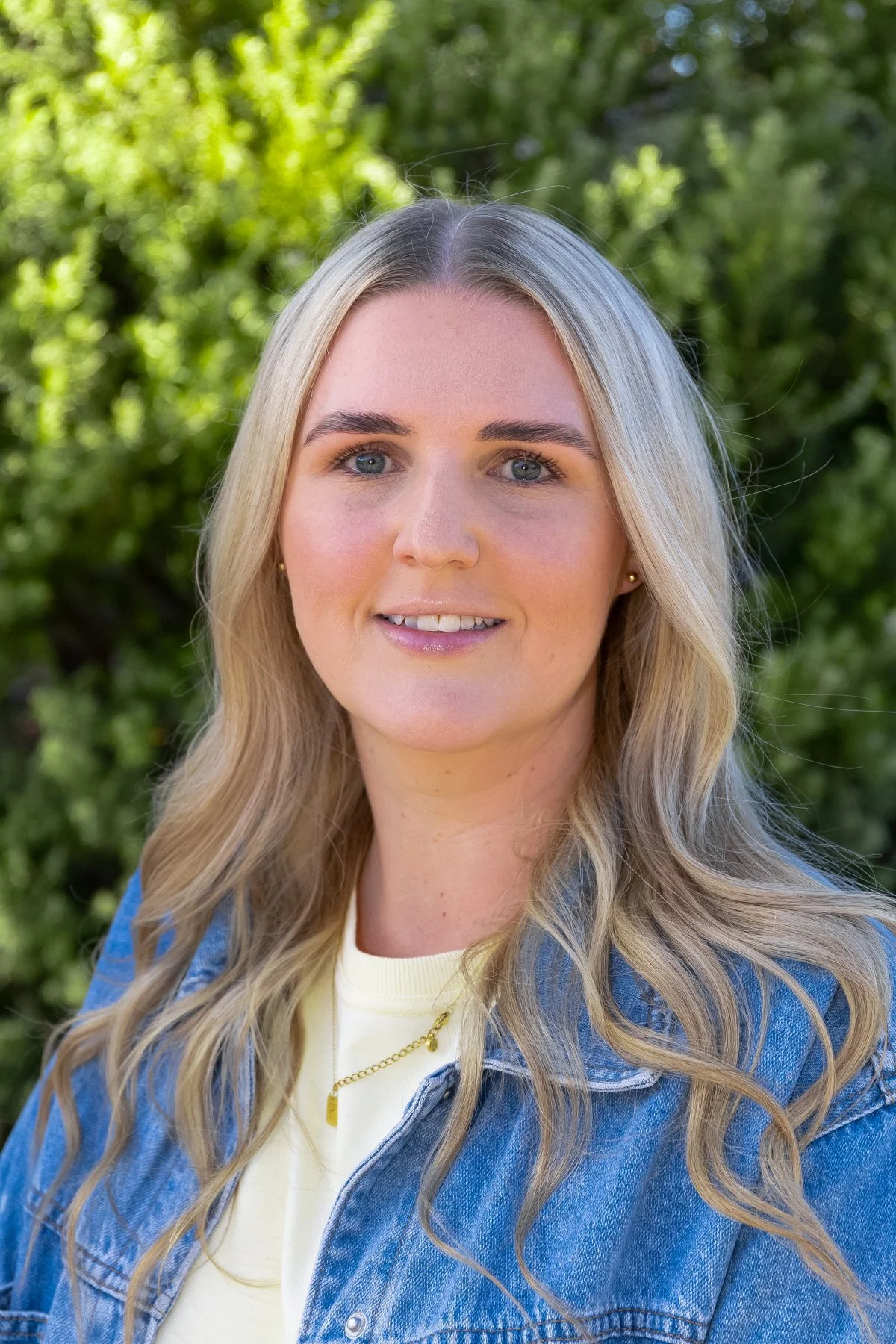 A young woman with long blonde hair and blue eyes smiling outdoors in front of green foliage, wearing a denim jacket and a light-colored shirt.