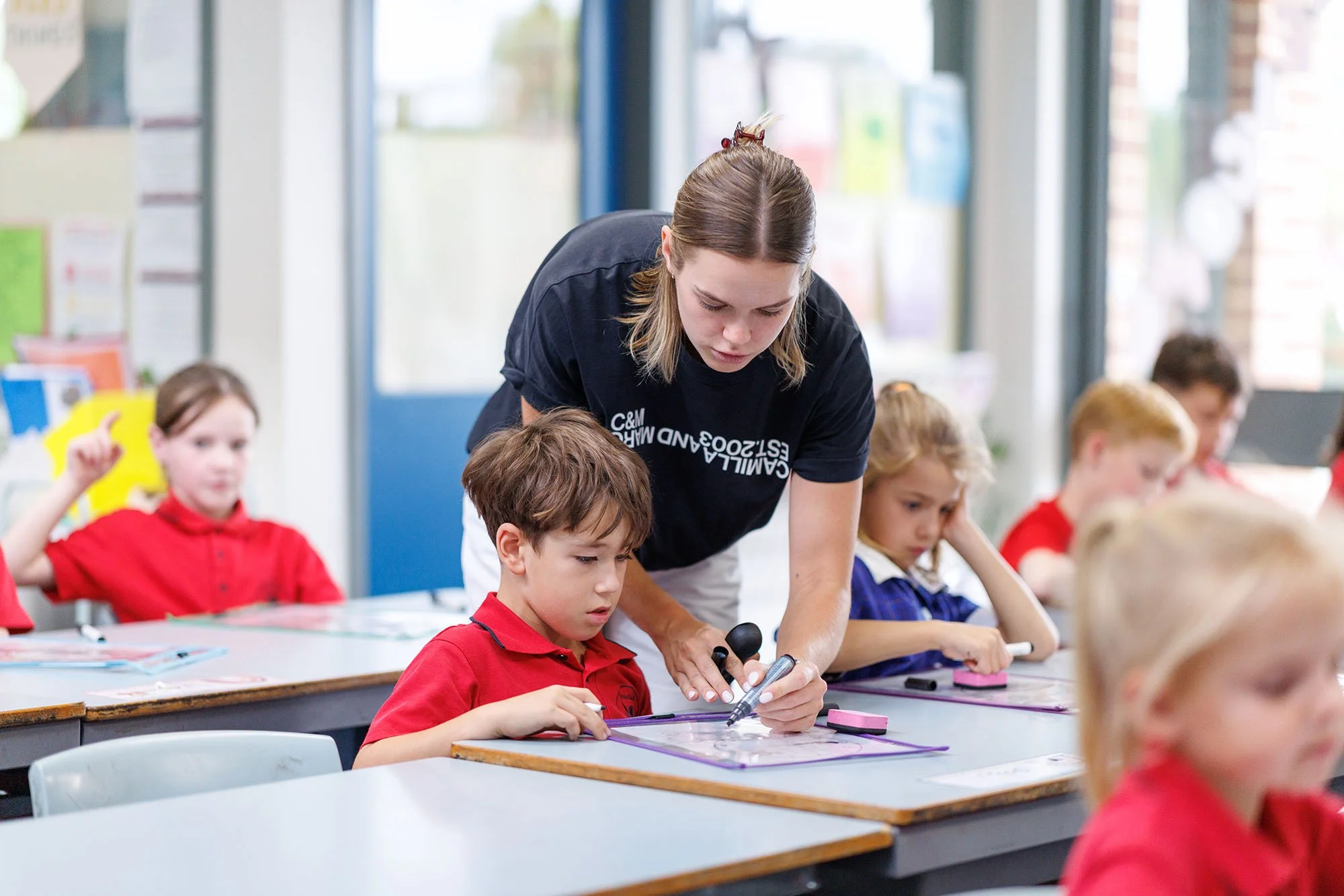 A classroom scene with a teacher instructing students seated at desks. The teacher is leaning over a student, showing him something on a transparent writing board, with other students visible in the background.