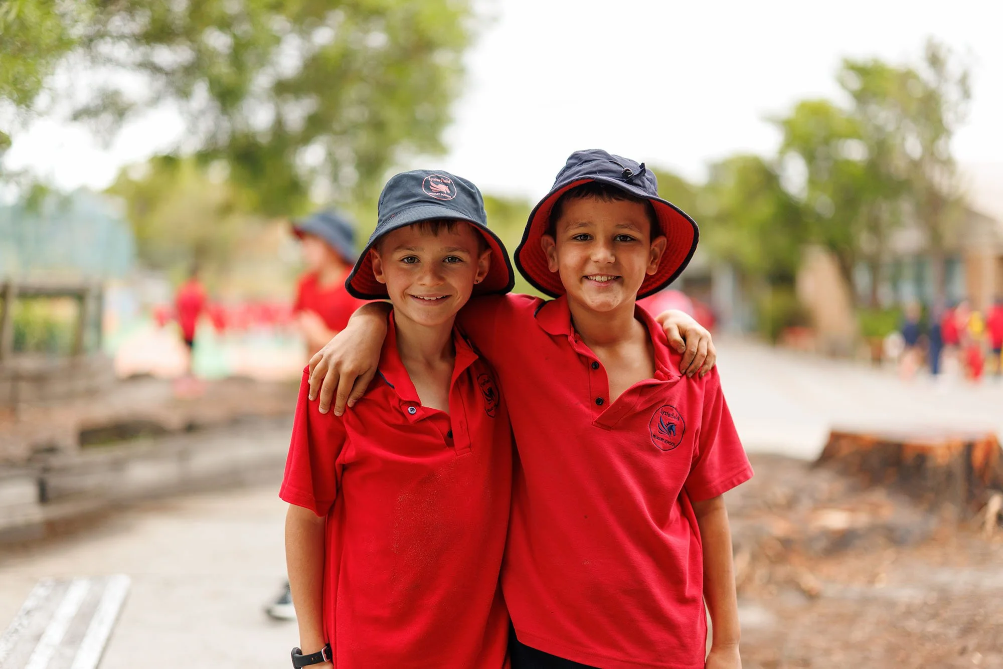 Two boys in red school uniforms and wide-brimmed hats smiling with their arms around each other outdoors.